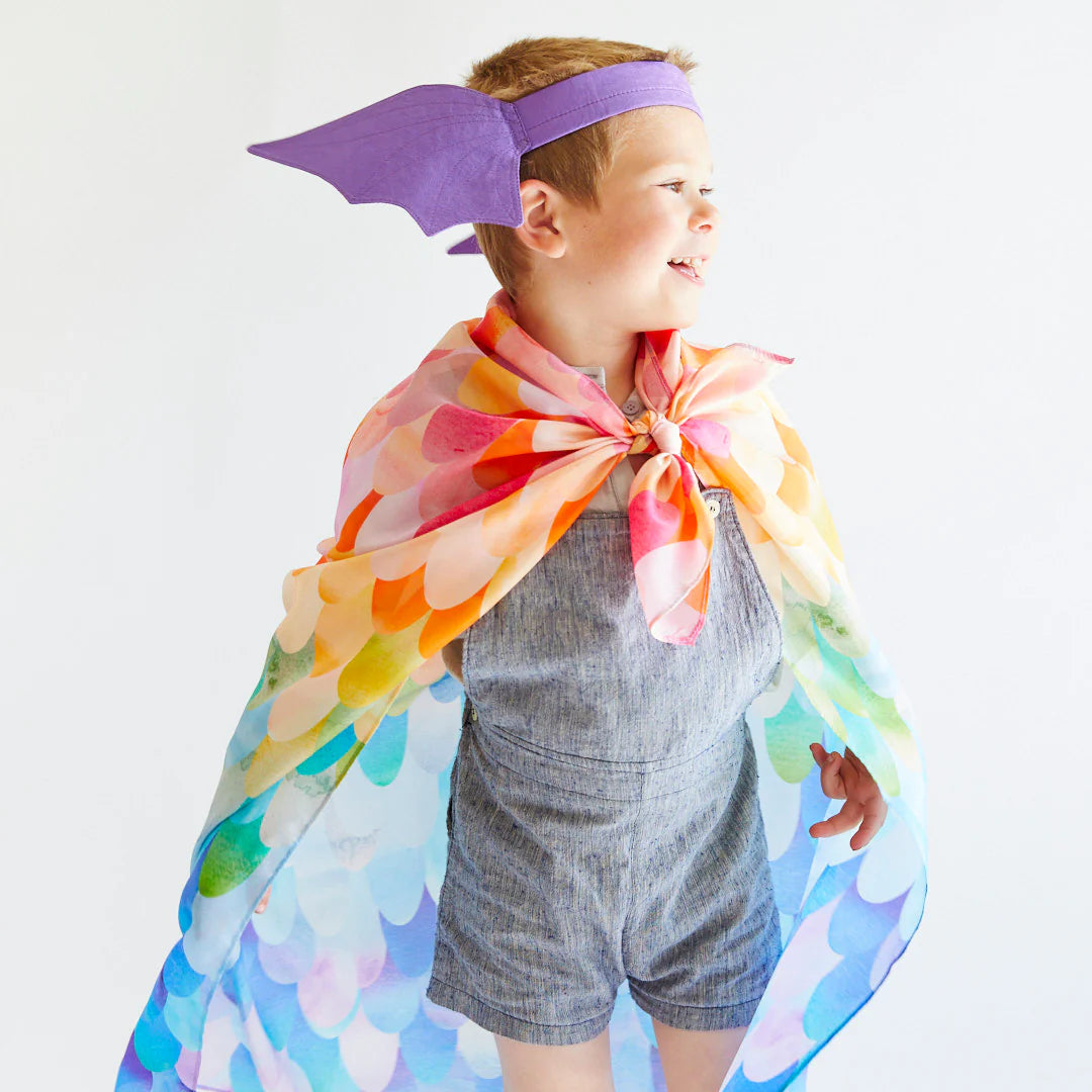 A smiling child engaged in dress-up play wears a vibrant cape with a rainbow scale pattern and the Dragon Ears Purple headband by SARAH'S SILKS, standing against a plain background. Sporting a gray romper, the child looks to the side, embodying the essence of open-ended play.