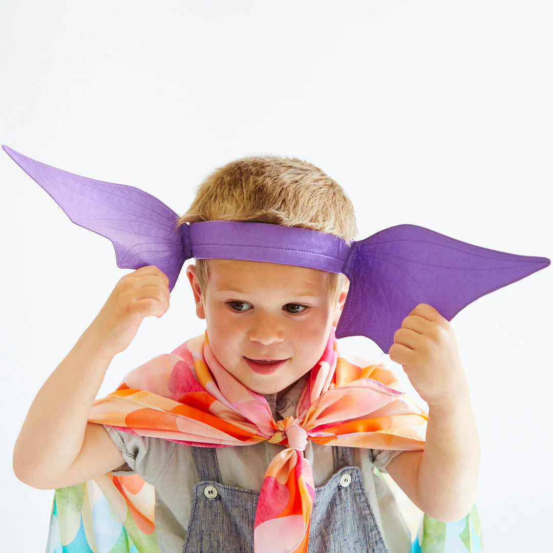 A young child with short hair wears the Dragon Ears Purple headband from SARAH'S SILKS, which resembles large bat-like ears. Dressed in denim overalls and a colorful, feather-patterned cape, they embody the spirit of imaginative play. Smiling as they look to the side against a plain white background, they showcase endless possibilities for open-ended play.