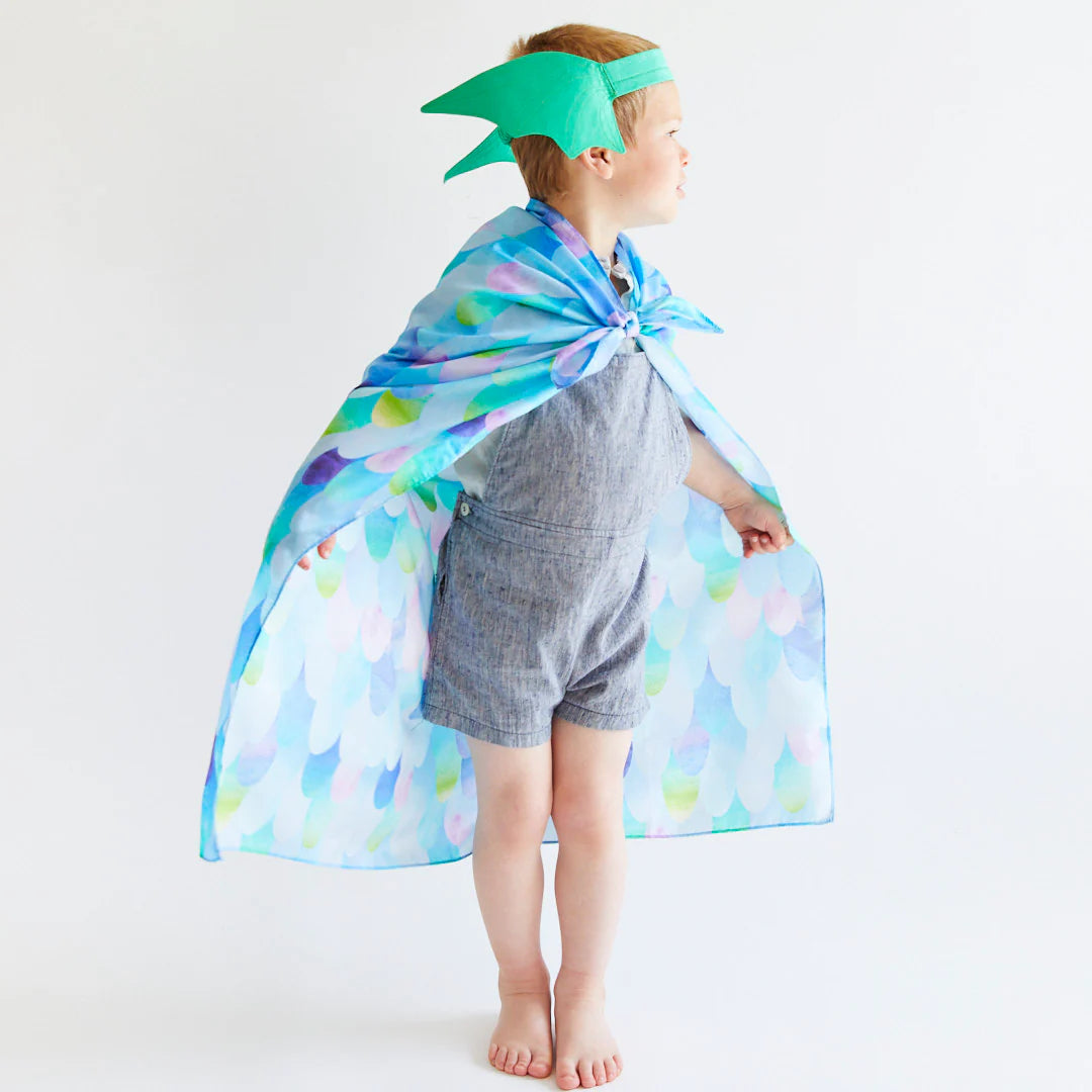 A child dressed in a light blue romper stands with arms akimbo, wearing SARAH'S SILKS' Blue Dragon Playsilk, adorned with multicolored scales and a green dragon headband. Embracing open-ended play, they are ready to explore the world of magical creatures against a plain white backdrop.