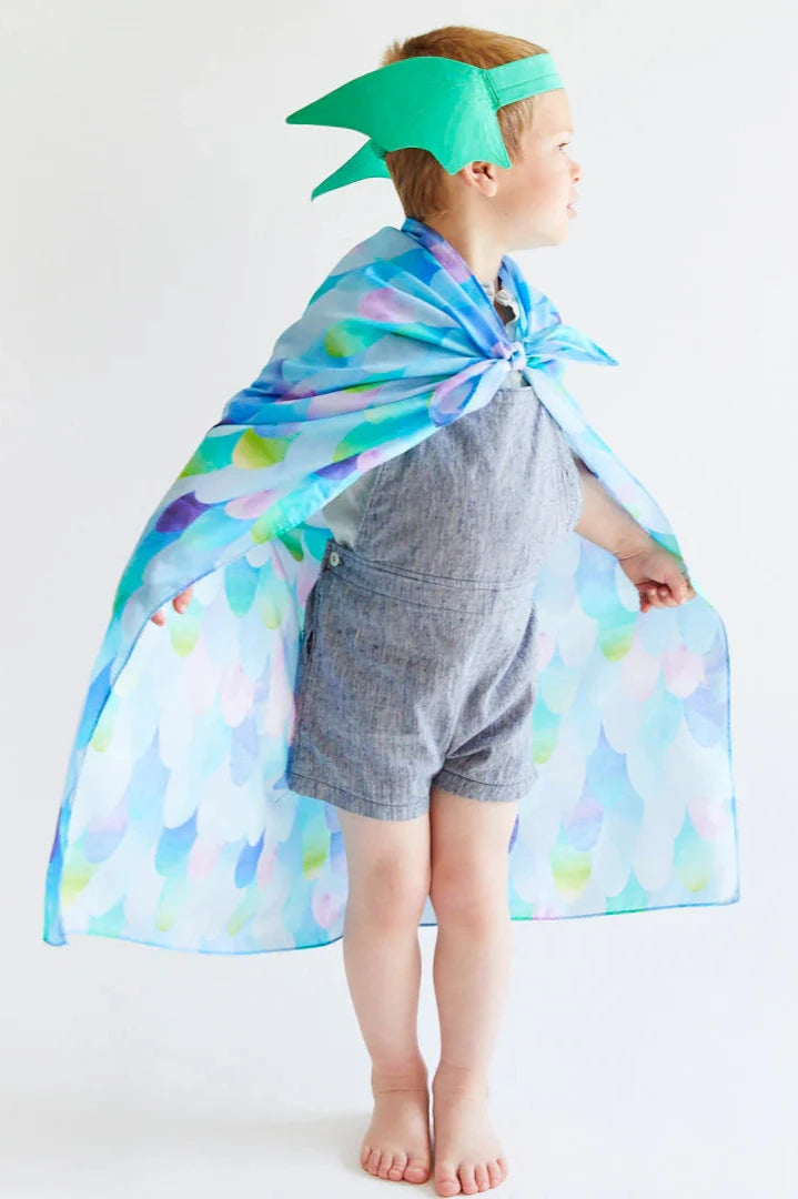 A young child, immersed in dress-up play, sports the Dragon Ears Green headband from SARAH'S SILKS along with a vibrant, scale-patterned cape over a blue romper, gazing to the side against a plain background.