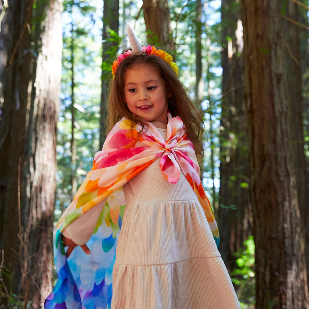 In the heart of the enchanting forest, a young child wears a cream dress and a SARAH'S SILKS Rainbow Unicorn Headband, ideal for captivating dramatic play. Sunlight streams through the tall trees, creating an ethereal glow that amplifies the magical ambiance of this whimsical dress-up scene.