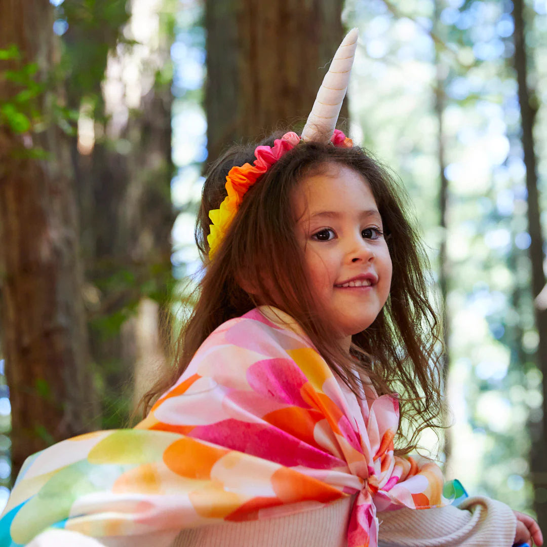 A young child with long hair happily wears a vibrant Rainbow Unicorn Headband from SARAH'S SILKS and a colorful cape. While engaging in dress-up play, they enjoy the enchanting forest setting, surrounded by towering trees with sunlight streaming through the leaves, exuding joy and beaming with happiness.