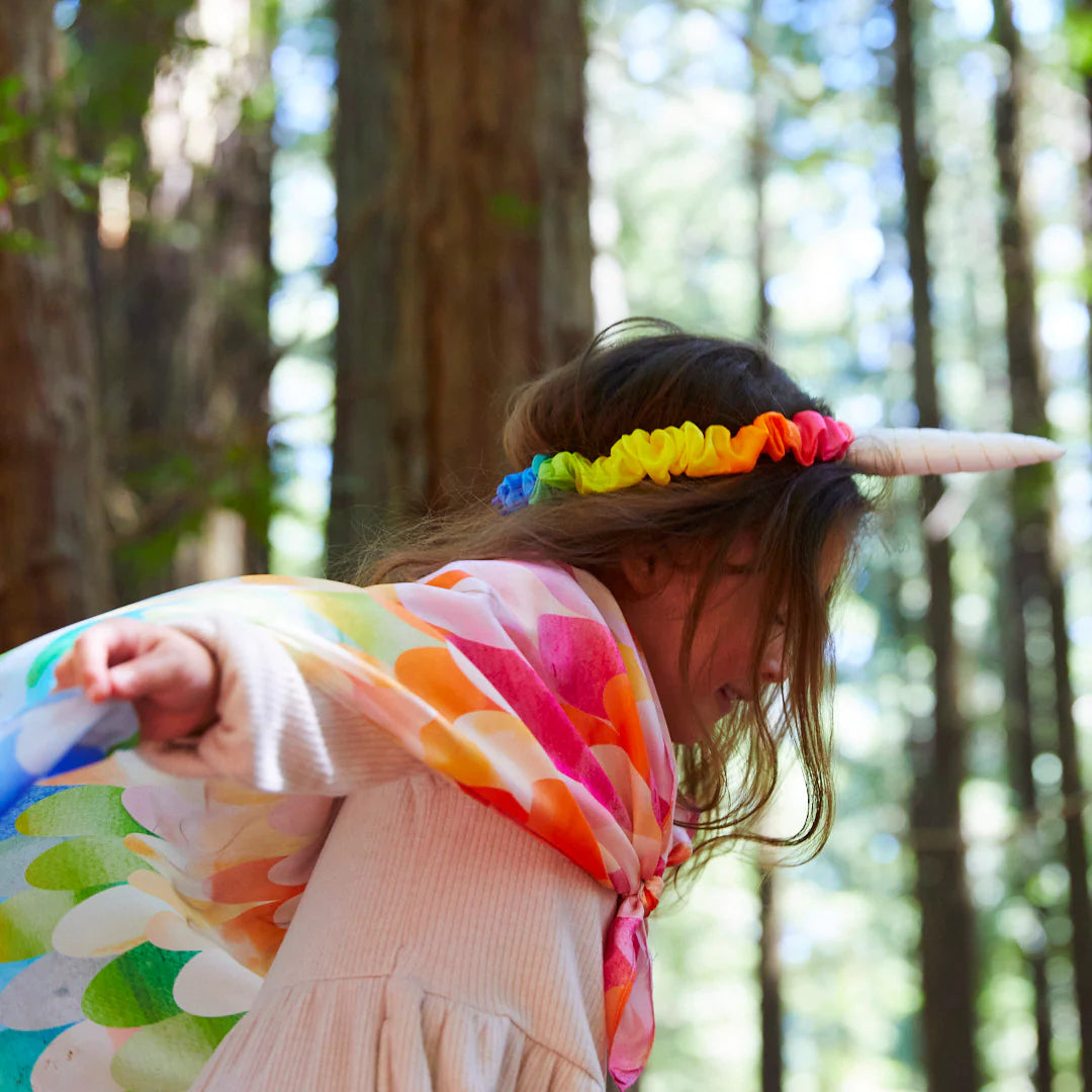 A child wearing SARAH'S SILKS Rainbow Unicorn Headband and a colorful cape plays in a forest. Tall trees surround them, while dappled sunlight filters through the leaves, creating a magical atmosphere perfect for dress-up play.