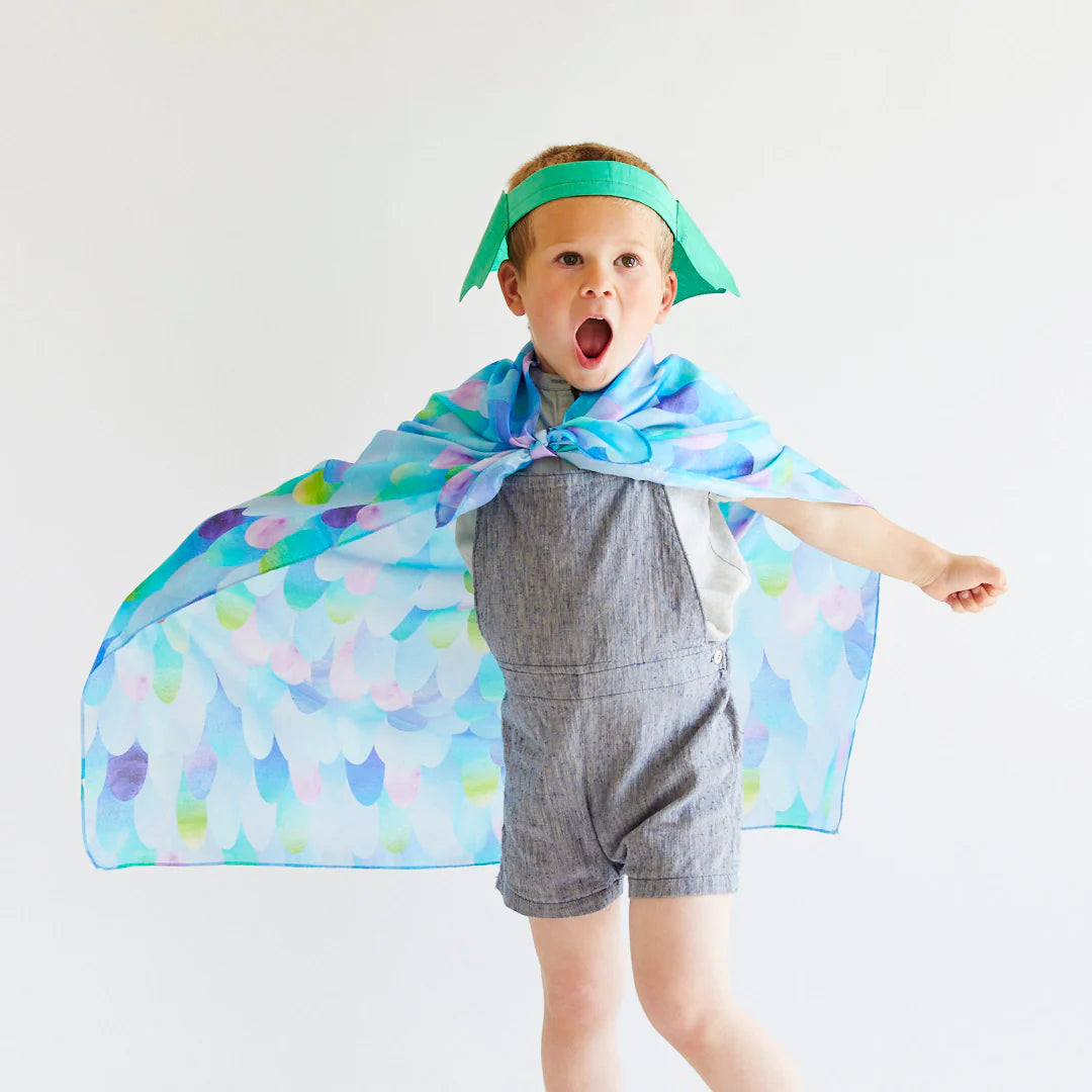 A child stands against a plain white background, adorned in a vibrant cape and a green paper crown. They embody the spirit of imaginative play, looking excited with an open mouth and one arm raised in a superhero pose, using their Blue Dragon Playsilk by SARAH'S SILKS.