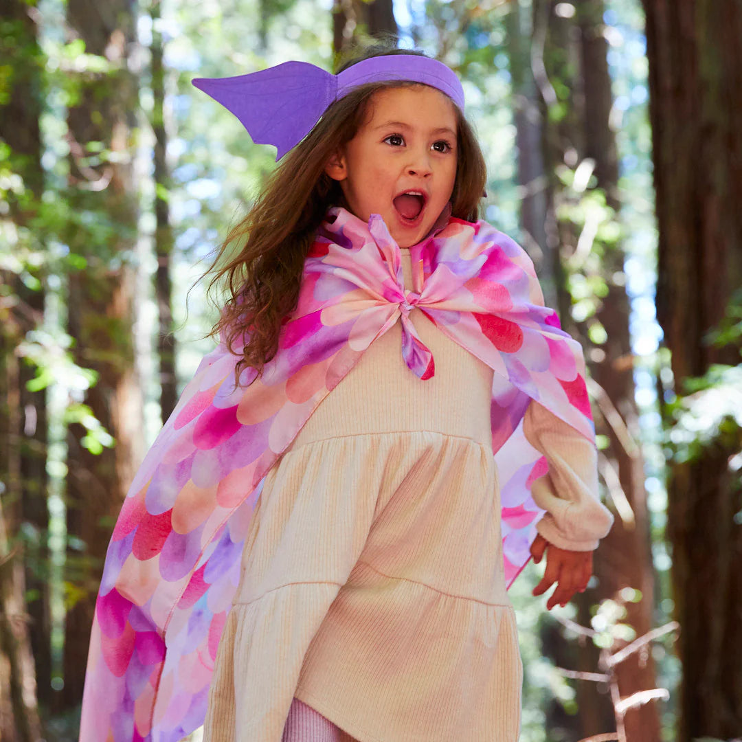 A young girl in a forest, dressed for dress-up play, is thrilled as she runs wearing SARAH'S SILKS' Dragon Ears Purple cape and matching headband. She is surrounded by tall trees and lush greenery, creating a magical atmosphere perfect for open-ended play.