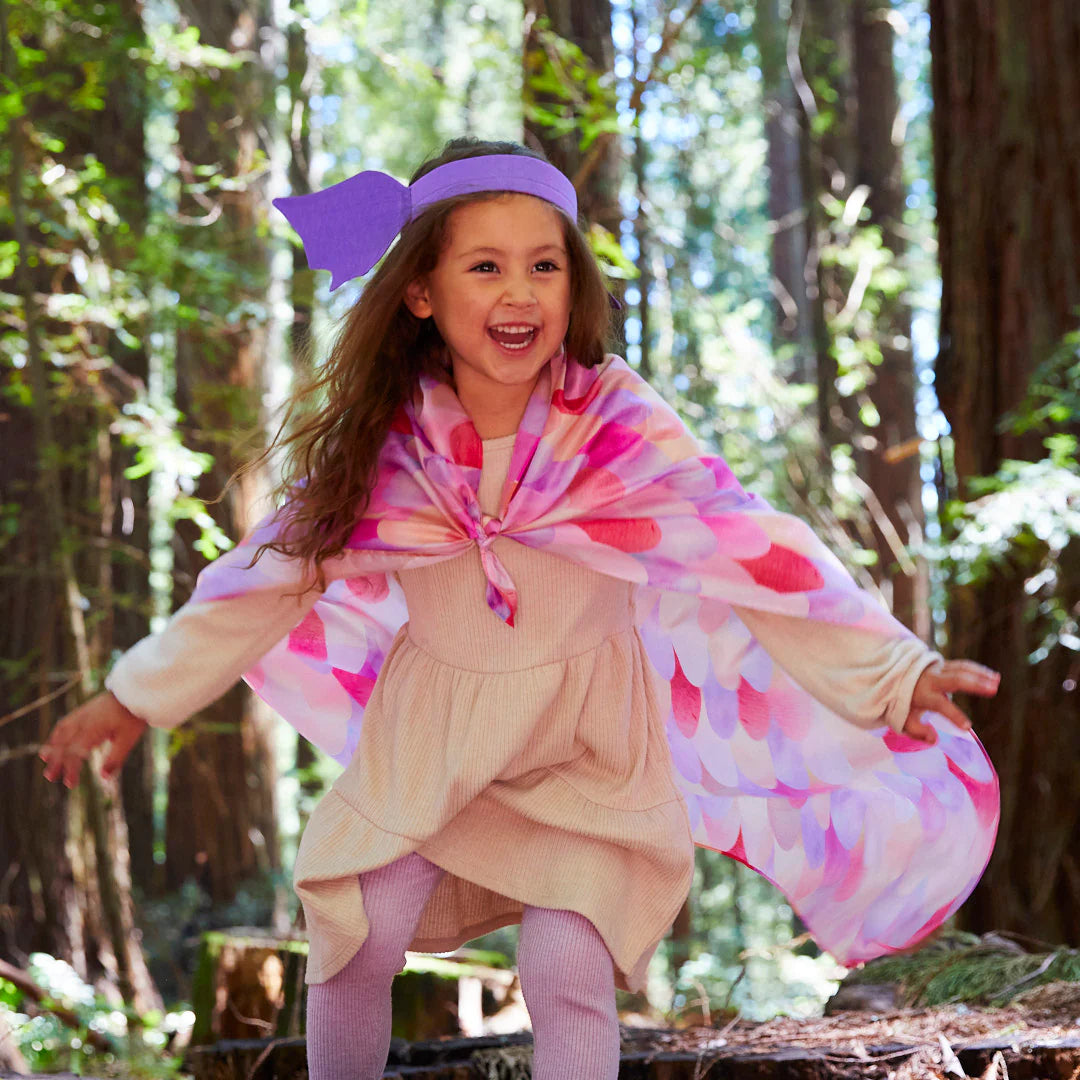 A joyful young girl with a purple headband and a colorful cape from SARAH'S SILKS, adorned with the Pink Unicorn Playsilk, runs through the forest. She wears a light dress and leggings, embracing open-ended play while dappled sunlight filters through the towering trees.