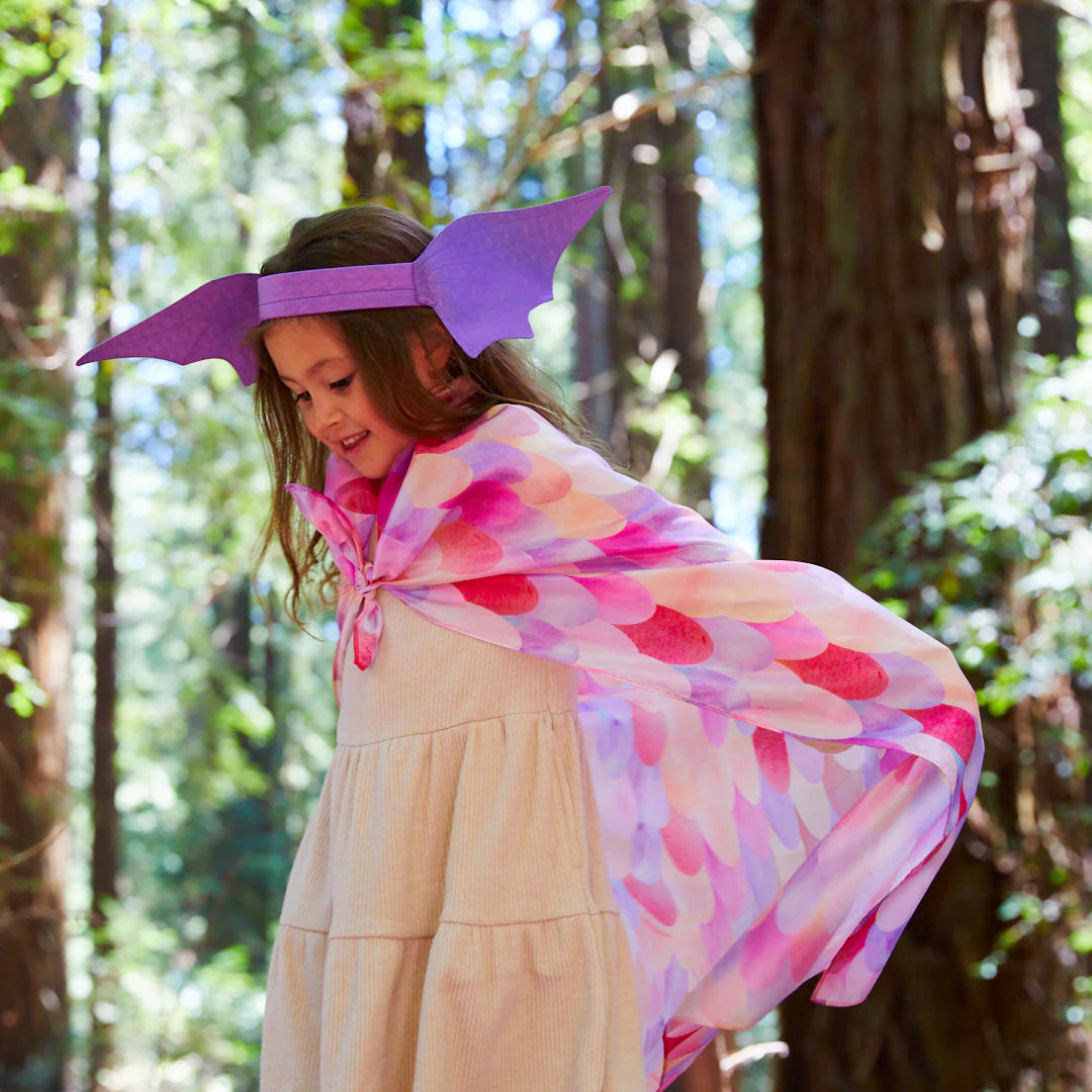 A young girl in a forest is dressed in the enchanting Dragon Ears Purple costume by SARAH'S SILKS, featuring a vibrant scale-patterned cape and dragon ears headband. With a joyful smile, she gazes downwards, surrounded by towering trees and lush greenery, fully immersed in her imaginative dress-up adventure.