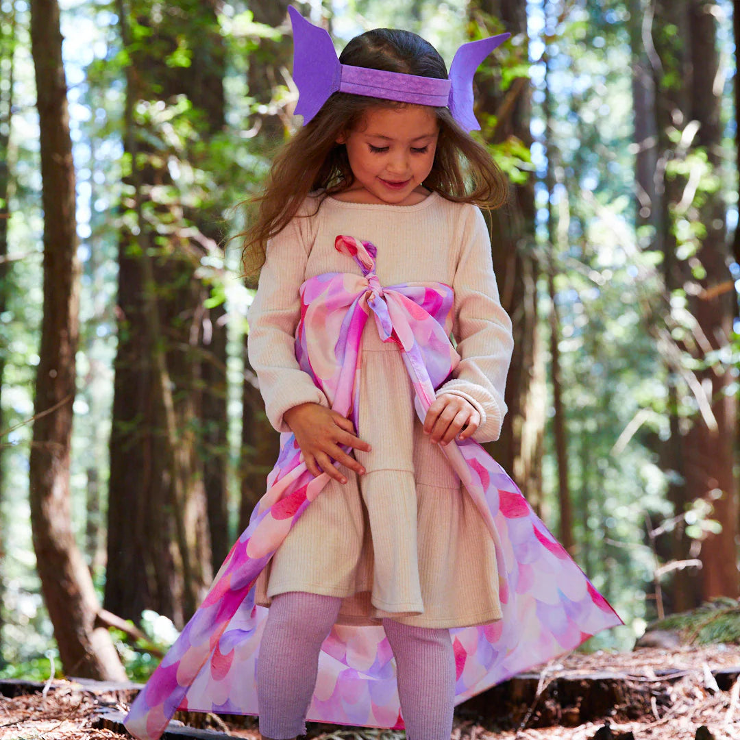 A young girl stands in a forest, fully engaged in dress-up play. She is wearing a light-colored dress paired with a cape featuring pink and purple petal patterns and a paper crown decorated with Dragon Ears Purple from SARAH'S SILKS on her head. Smiling as she looks down, she is enveloped by tall trees, sparking imaginative play adventures.