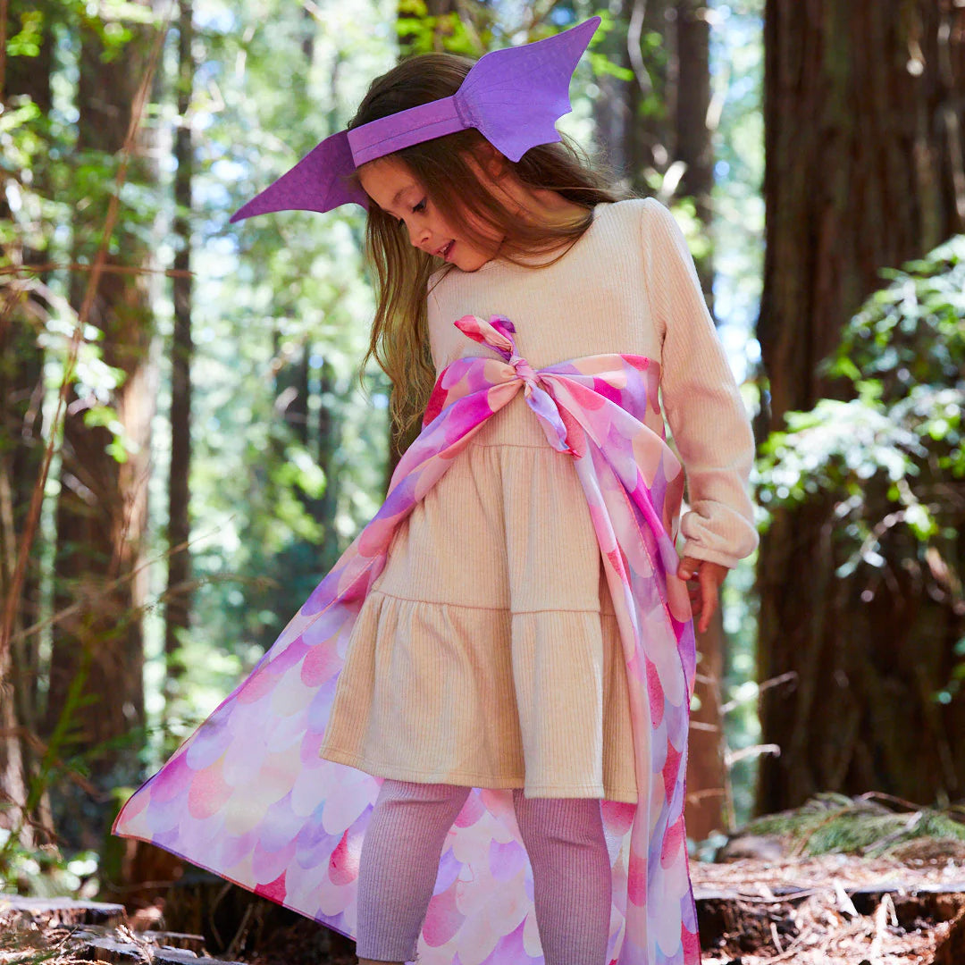 A young child in a beige dress and cape decorated with SARAH'S SILKS Pink Unicorn Playsilk stands in a forest. They wear a whimsical purple headband with large ears, perfect for open-ended play. Sunlight filters through the tall trees, casting a serene atmosphere reminiscent of magical settings from playsilks.