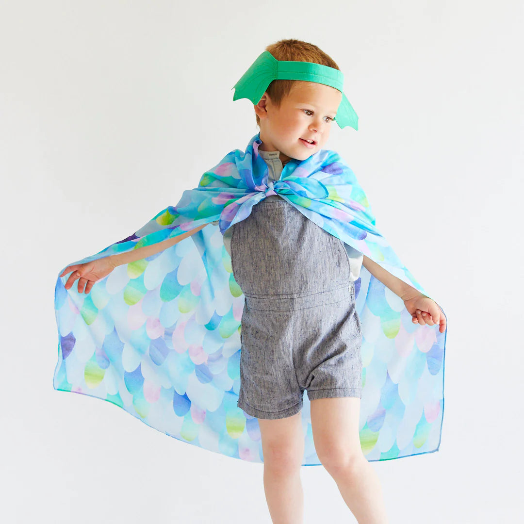 A creative child strikes a playful pose, draped in a vibrant, patterned cape and adorned with SARAH'S SILKS' Dragon Ears Green headband. They wear a basic gray romper against a simple background, capturing the essence of dress-up play.