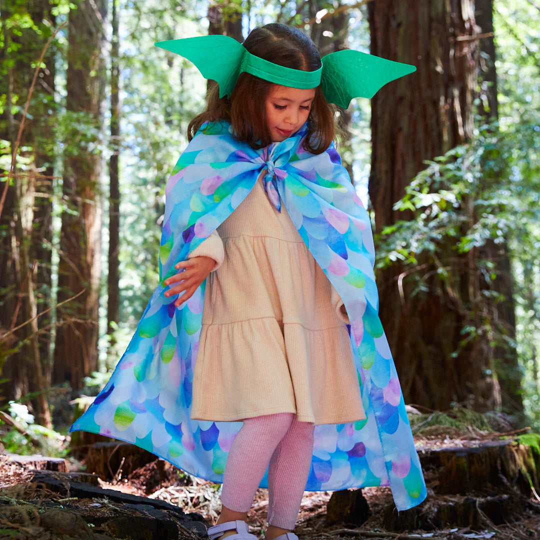 In a lush forest, a child dressed in a beige dress, pink leggings, and an array of colorful SARAH'S SILKS Playsilks along with Blue Dragon headgear stands. Sunlight filters through the tall trees, illuminating this enchanting scene of open-ended play.