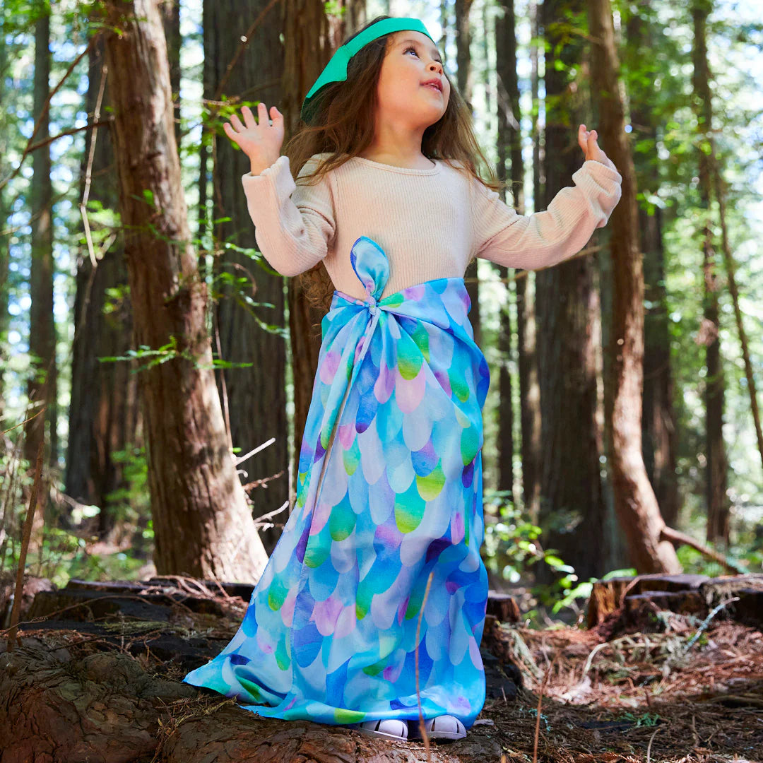 A young child stands on the forest floor, wearing a colorful, flowing skirt crafted from 100% pure silk and a matching headband. Their long brown hair cascades down as they gesture expressively with both hands. The tall trees and sunlight filtering through the leaves hint at the presence of the enchanting Blue Dragon Playsilk by SARAH'S SILKS in this magical scene.