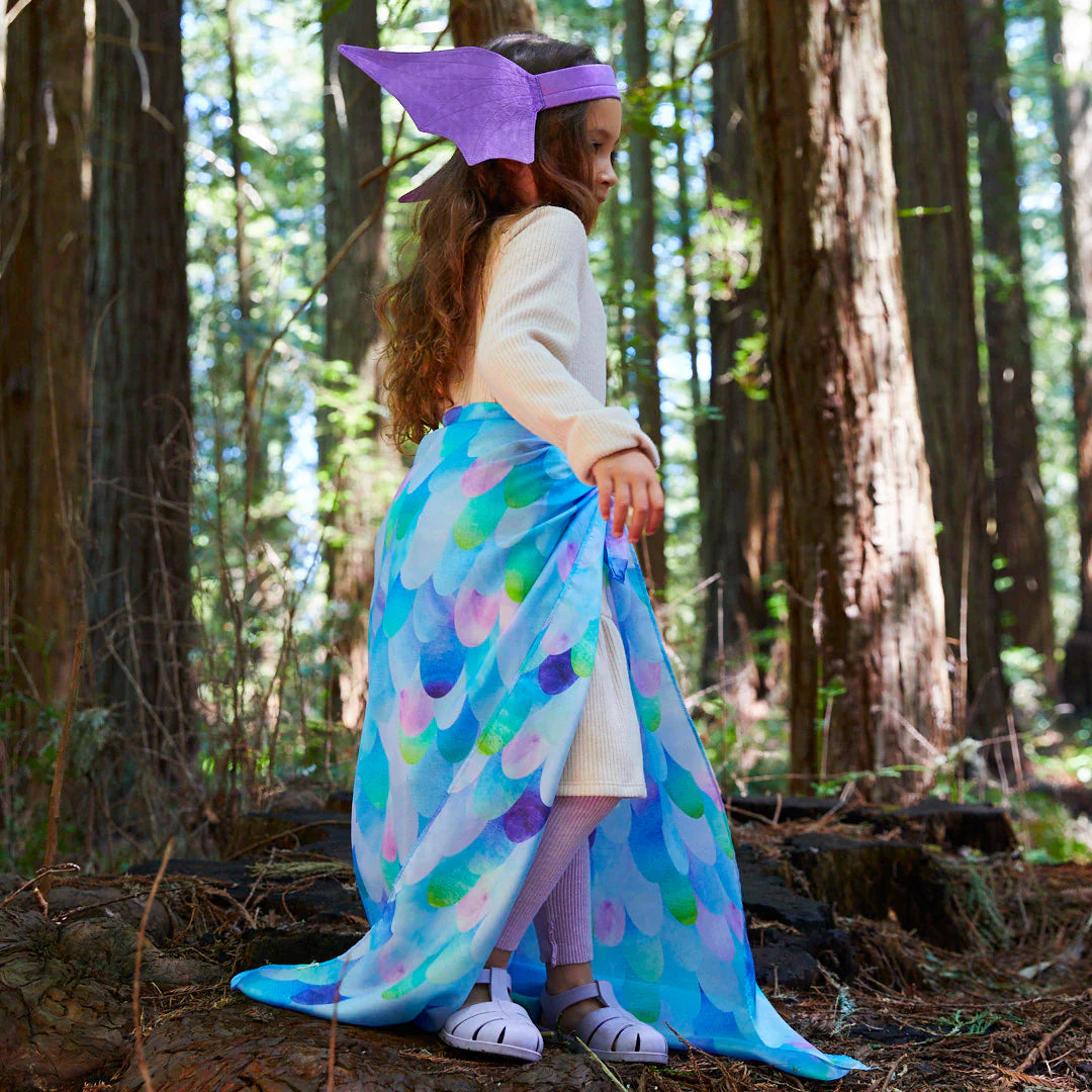 In a serene forest setting, a child dons an enchanting costume crafted by SARAH'S SILKS, featuring the Blue Dragon Playsilk. The whimsical attire includes a purple headpiece and a silk cape that beautifully mimics fish scales. The tall trees and gentle natural light enhance the invitation for open-ended play in this magical atmosphere.