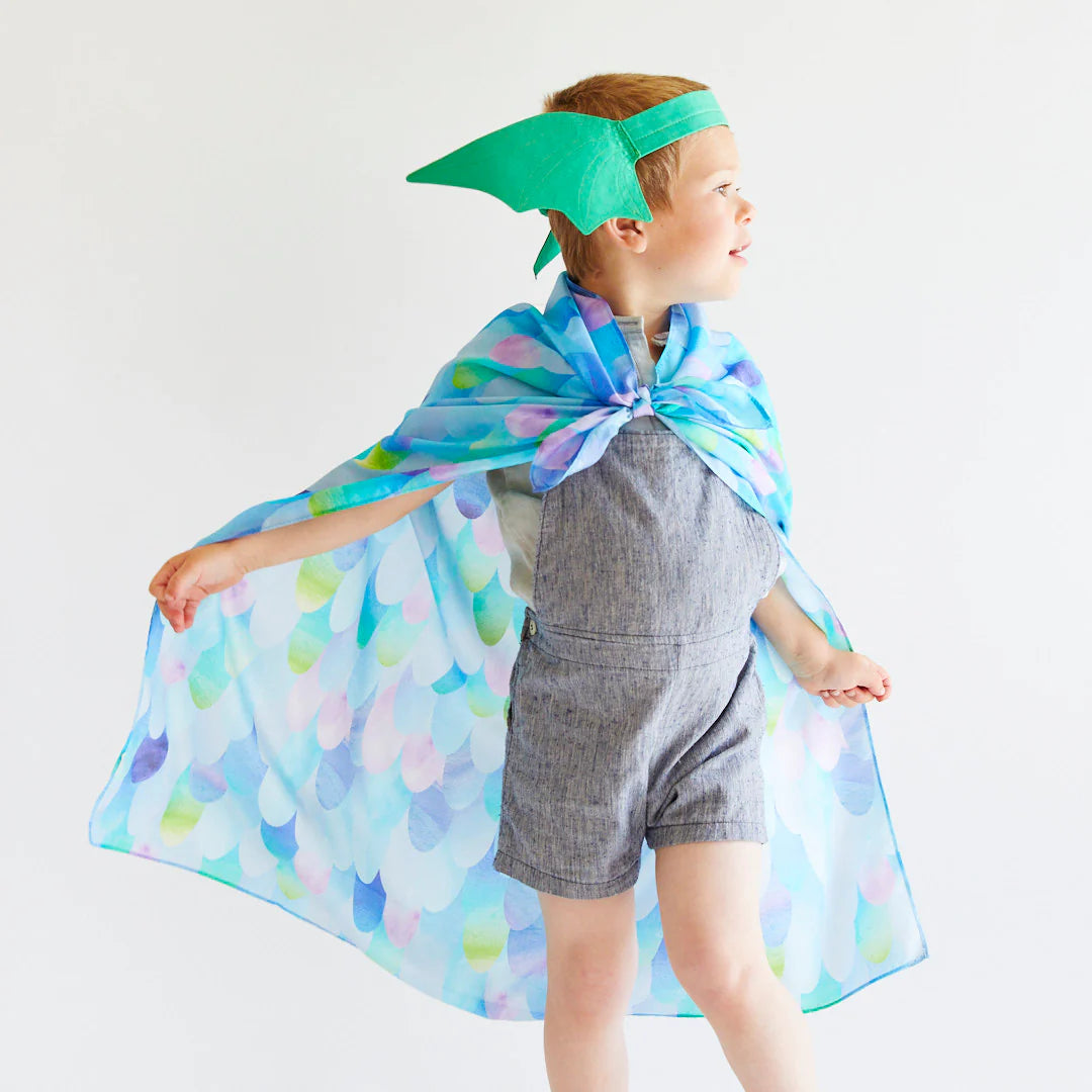 A child wearing a blue and purple textured cape and the Dragon Ears Green headband by SARAH'S SILKS stands on a light background. This imaginative child, dressed in a gray romper, poses with arms slightly spread, looking to the side, embodying the spirit of dress-up play.