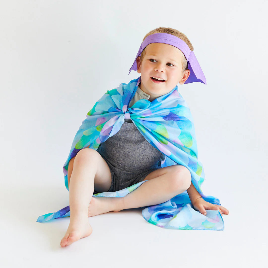 A young child sits on the floor wearing a Blue Dragon cape made of 100% pure silk and a purple headband with pointy ends. The child is smiling in a gray outfit, embodying SARAH'S SILKS Blue Dragon Playsilk in open-ended play against a plain white background.