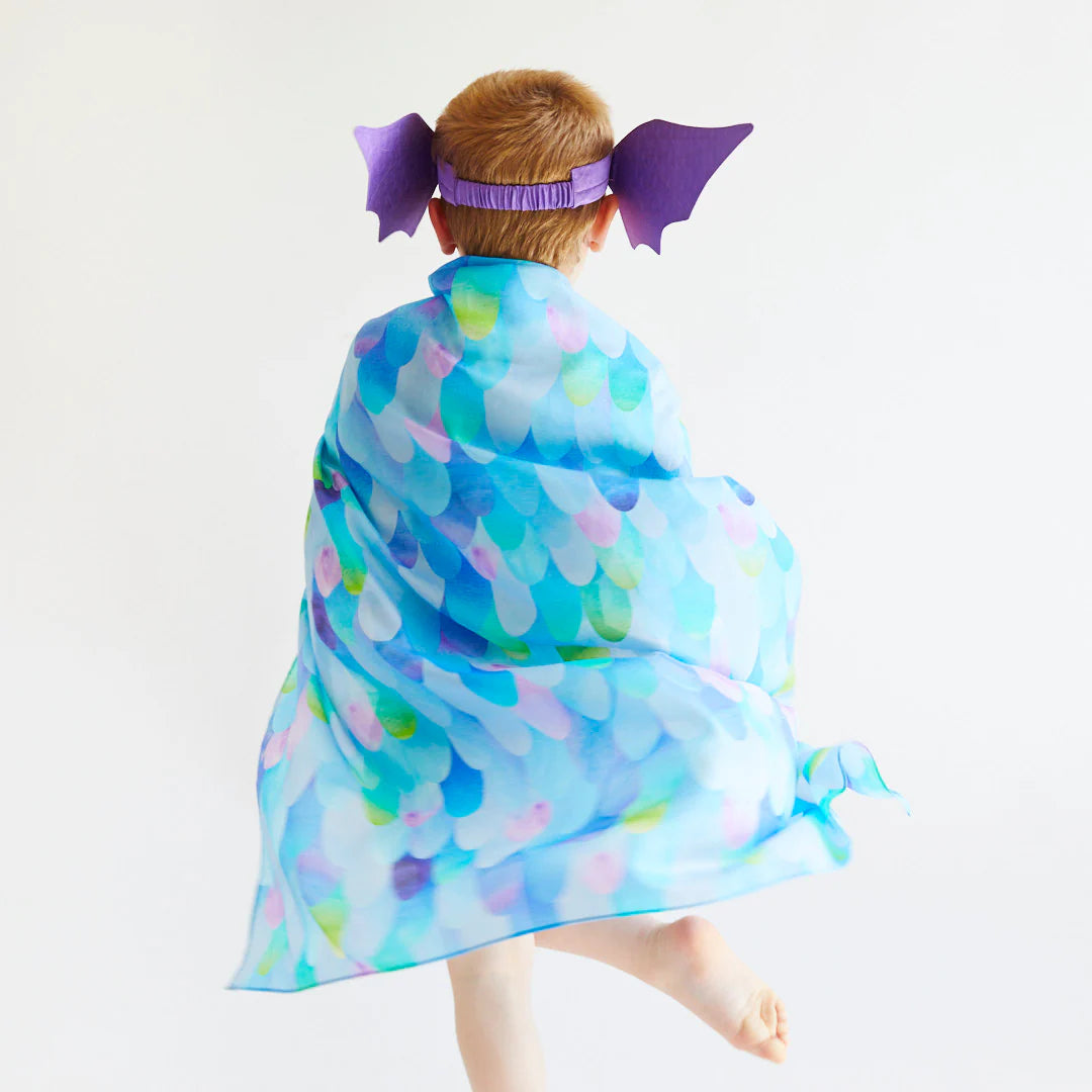 A child with short blonde hair joyfully leaps with one foot in the air, draped in a colorful, scale-patterned fabric from SARAH'S SILKS, made of 100% pure silk. The ensemble includes a purple headband featuring dragon wings—a delightful nod to the Blue Dragon Playsilk. All set against a white background.