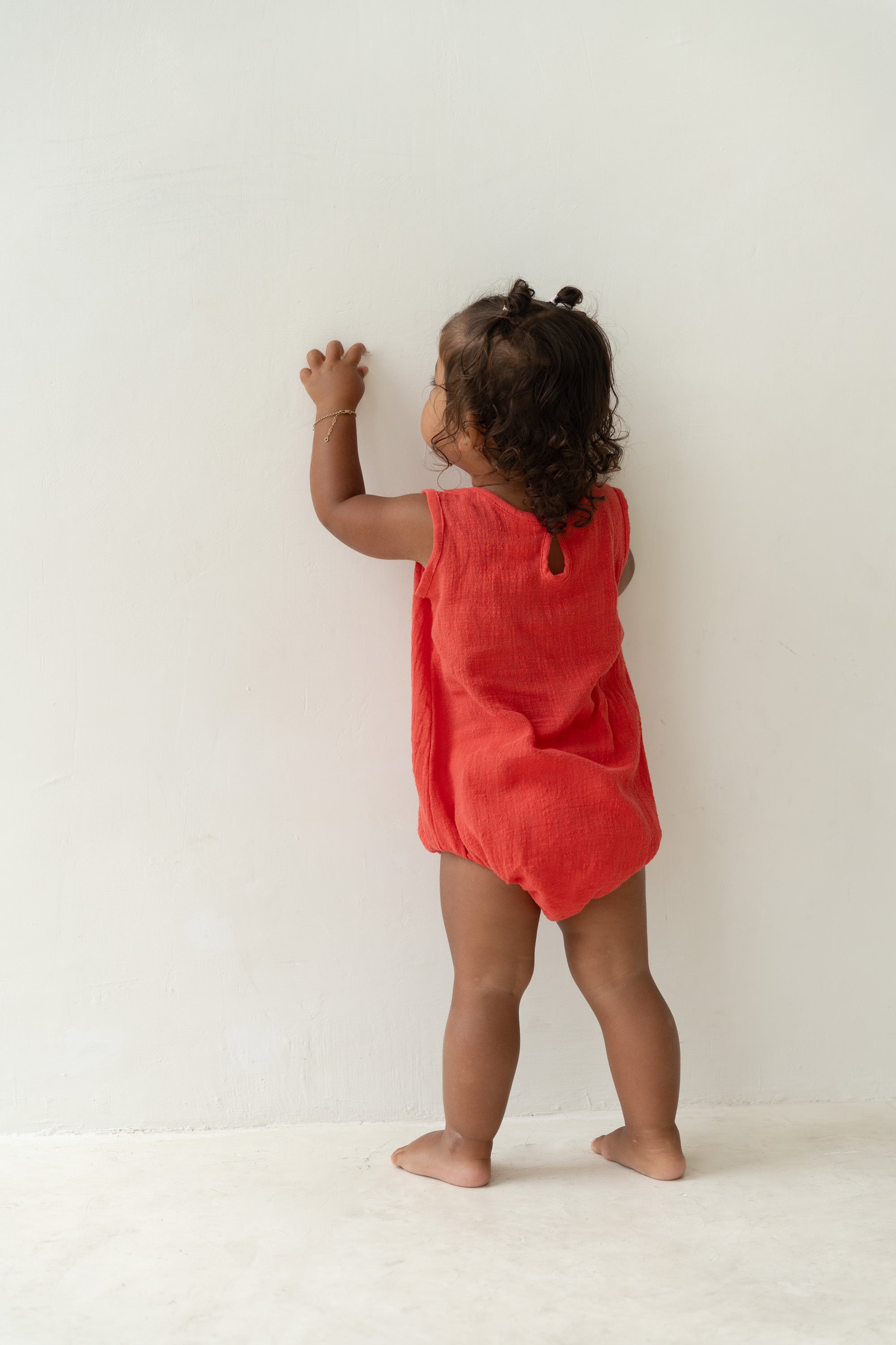 A curly-haired toddler stands barefoot on a light floor, reaching up to a white wall, wearing ILLOURA THE LABEL’s Mars Romper Watermelon with coconut buttons.