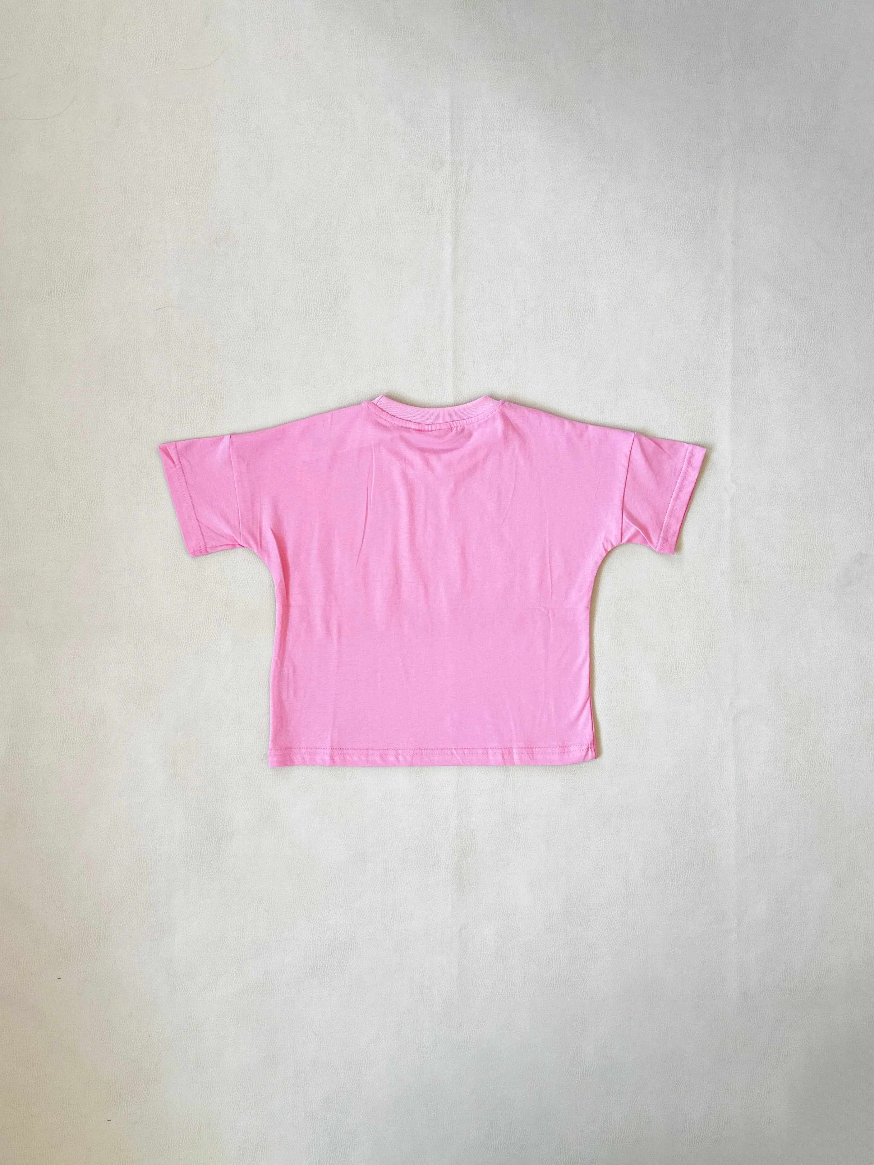 The Tiny Trove ~ Farmer's Market Relaxed Tee Bubblegum by TINY TROVE, made of soft cotton, is displayed on a light surface and viewed from the back. Its relaxed fit ensures comfort and style.