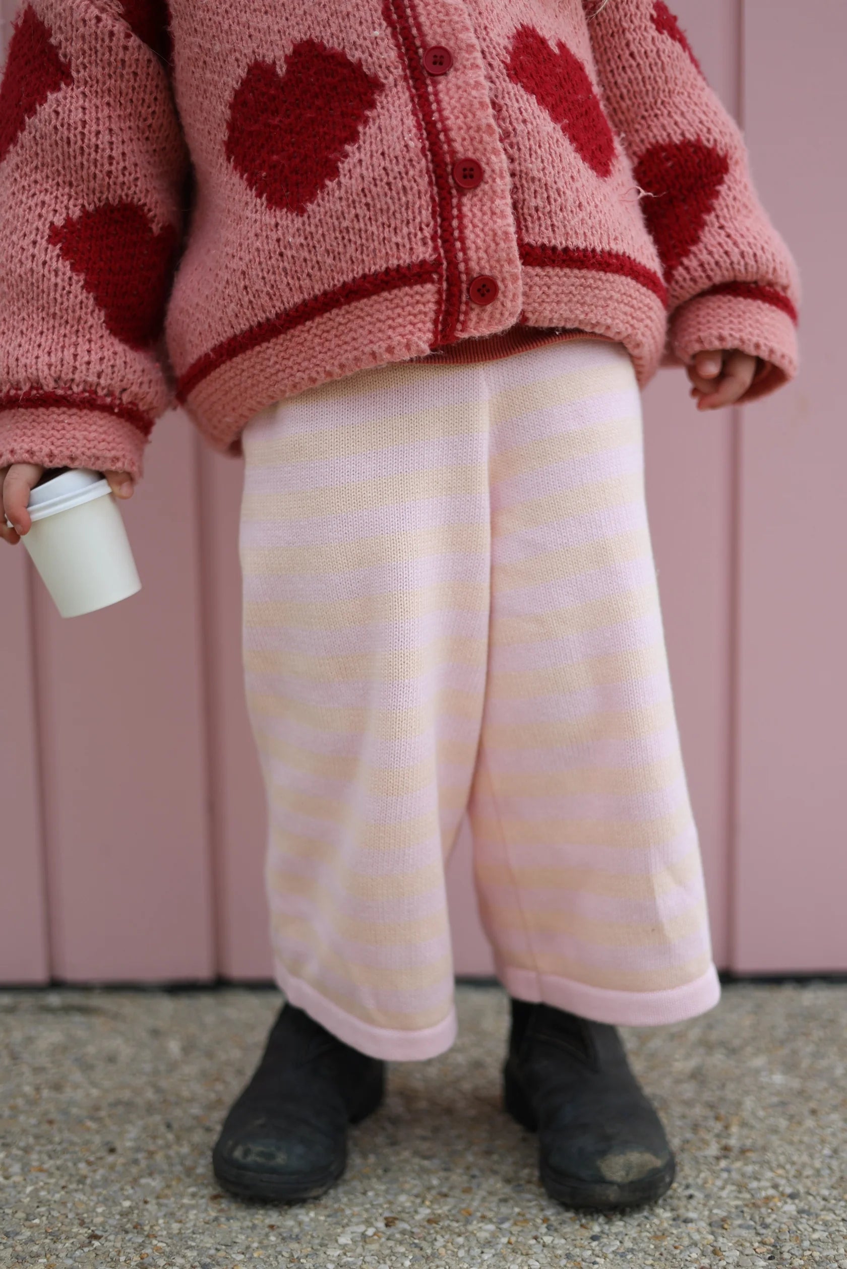 A child is standing in front of a pink wall, wearing a 100% cotton knit pink sweater adorned with red hearts and light pink striped Staple Pant Pink/Peach by MILKY DESIGNS, featuring an elastic waistband. They are also wearing dark shoes and holding a small white cup. Available sizing ranges from 0-4 years. The child's face is not visible in the image.