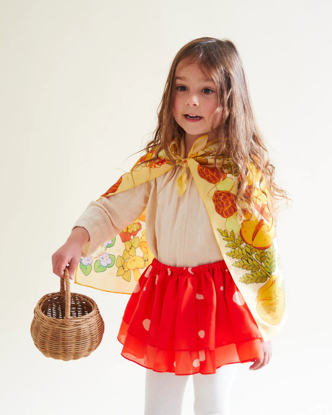 A young girl wearing a Mushroom Seek & Find Playsilk by SARAH'S SILKS, adorned with vibrant fruit patterns, stands against a light-colored background. Her outfit—a beige blouse paired with a red ruffled skirt featuring white polka dots and white tights—creates an ensemble perfect for whimsical nature walks. She holds a small wicker basket and smiles slightly.