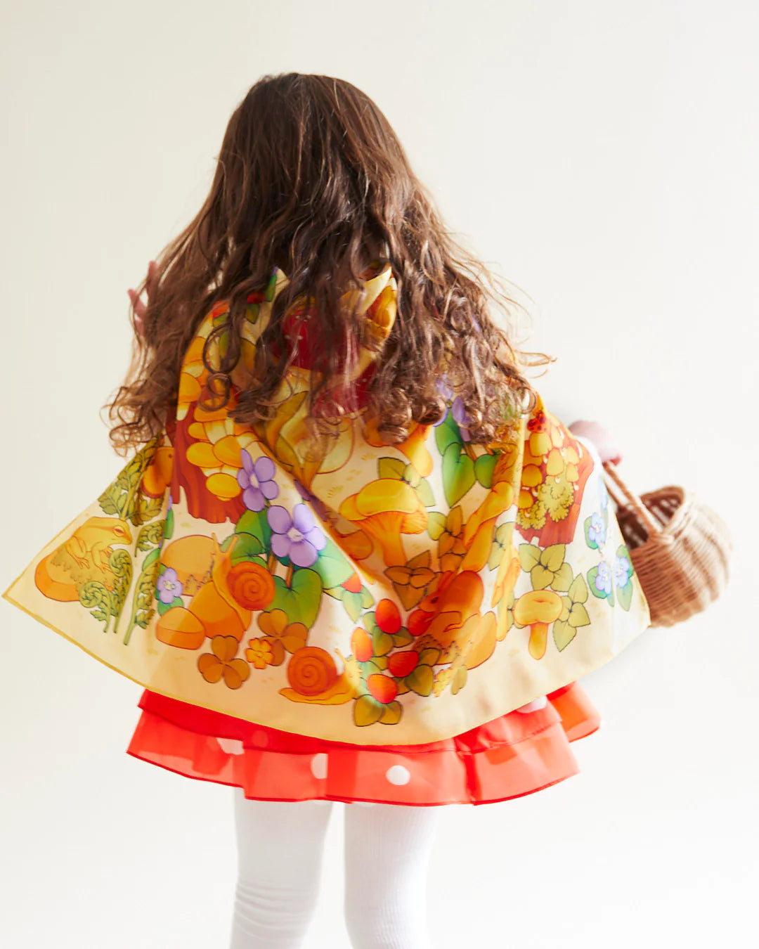 A girl with long hair stands facing away, wearing a vibrant floral scarf made from the Mushroom Seek & Find Playsilk by SARAH'S SILKS and a red dress featuring a white polka dot tiered skirt. She holds a woven basket, ready for nature walks, against a plain white background.