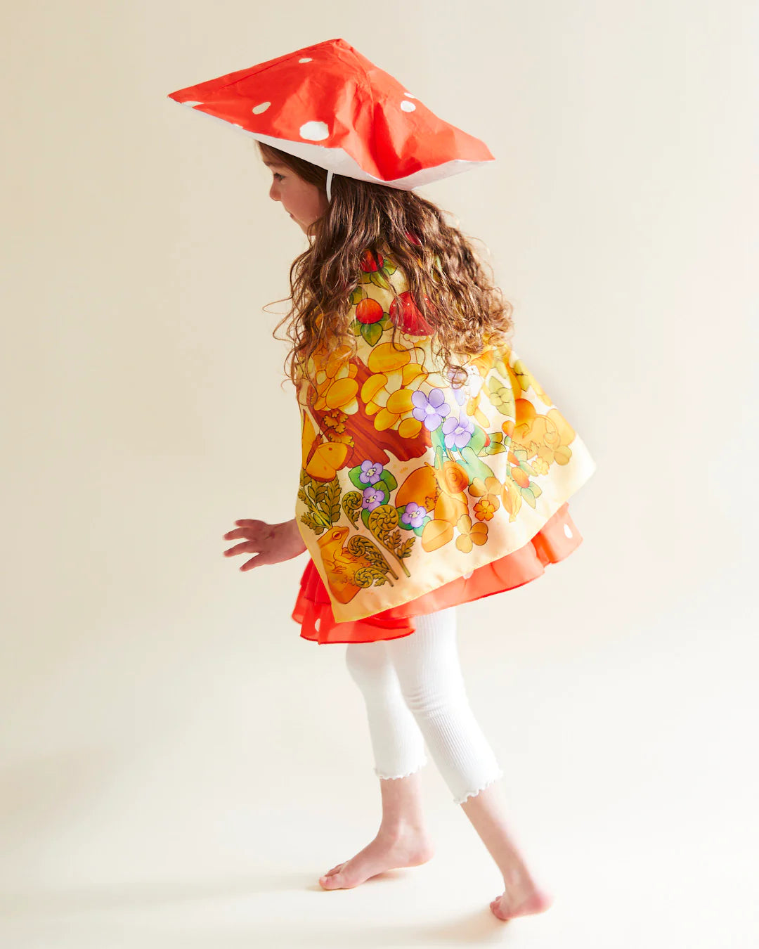 A child with long hair dons a large red mushroom-shaped hat with white spots and a colorful floral Mushroom Seek & Find Playsilk cape. Barefoot in white leggings, they wander against a plain background, reminiscent of whimsical nature walks. The cape is crafted from mulberry silk by SARAH'S SILKS.