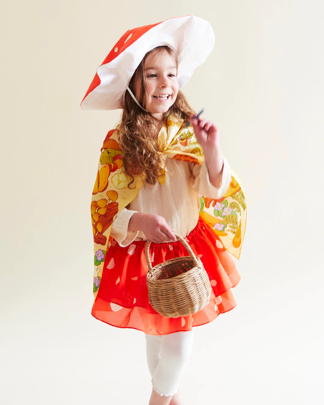 A young child dressed in a vibrant outfit featuring SARAH'S SILKS' Mushroom Seek & Find Playsilk, complete with a mushroom-themed hat and cape, holds a wicker basket. The ensemble, which includes a red skirt and white leggings crafted from luxurious mulberry silk, highlights their joy against a neutral background.