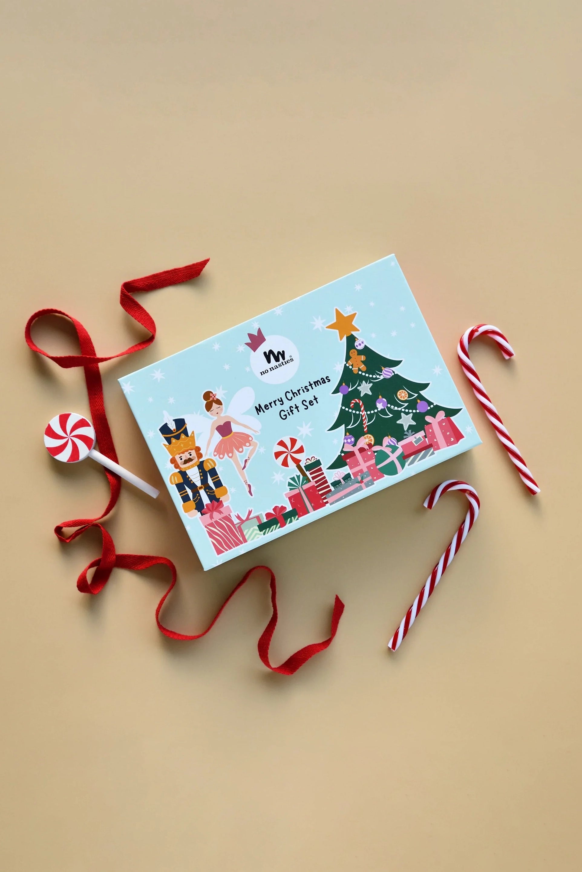 The No Nasties Signature Christmas Pop-Up Kids Makeup Box from NO NASTIES showcases festive illustrations, including a tree, presents, and whimsical pop-up holiday characters. It is decorated with red ribbon, candy canes, and a lollipop on a beige background.