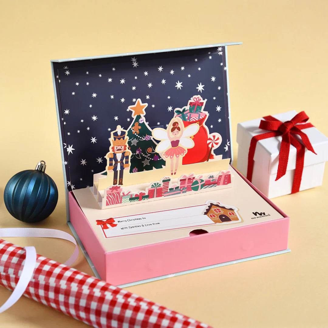 The No Nasties Signature Christmas Pop-Up Kids Makeup Box features whimsical Christmas characters such as a nutcracker and fairy, all housed in a pink box that resembles a children's makeup set. Nearby, a wrapped gift adorned with a red bow, blue ornament, gingham paper, and roll of tape completes this charming holiday scene.