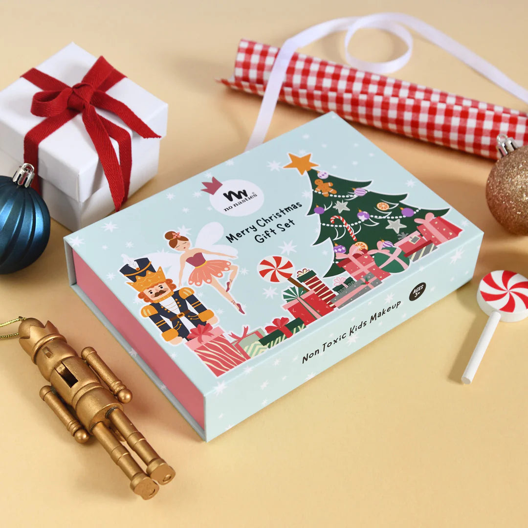 A festive scene showcases the NO NASTIES Signature Christmas Pop-Up Kids Makeup Box, adorned with Christmas characters and merry illustrations of nutcrackers and a tree. Surrounding this enchanting gift set are bauble ornaments, a wooden toy, a lollipop, and a wrapped present topped with a red bow, all contributing to the holiday magic.