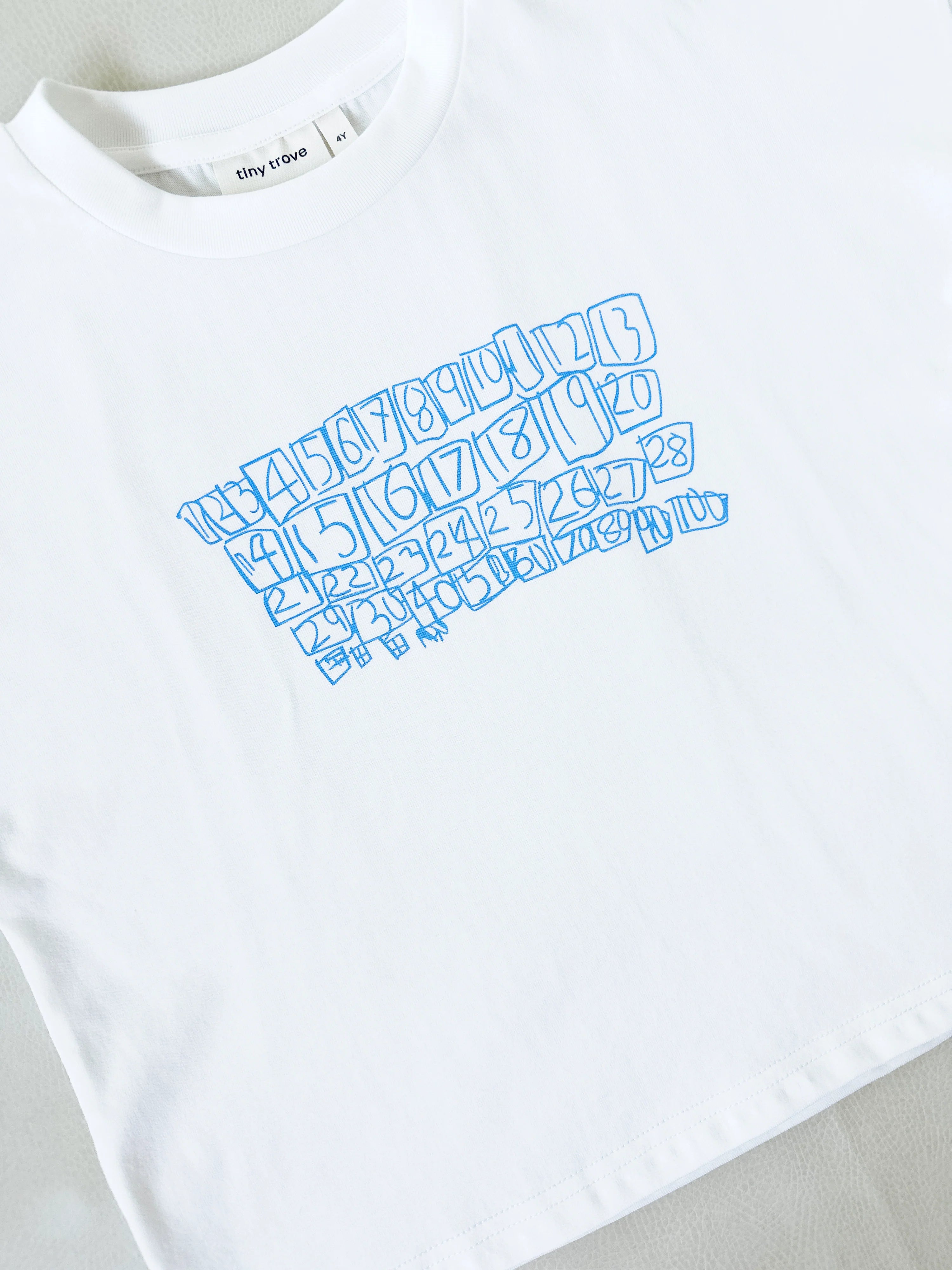 The TINY TROVE Parker Numbers Relaxed Tee White/Blue is an oversized 100% cotton kids’ tee with a playful blue hand-drawn numbers print and a signature “tiny trove” tag.