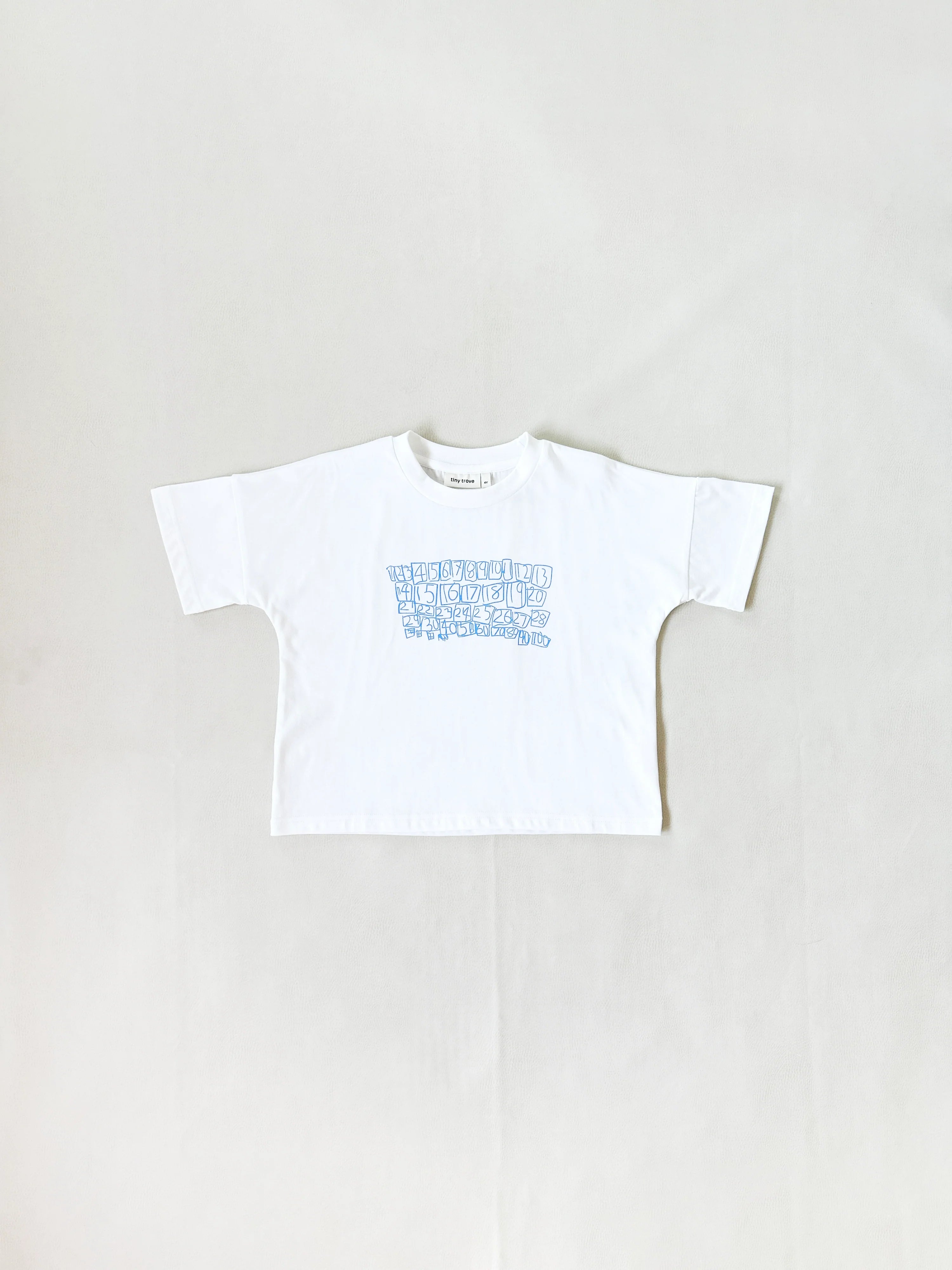 The Tiny Trove - Parker Numbers Relaxed Tee White/Blue by TINY TROVE is a kids' oversized t-shirt in white, made from 100% cotton jersey, with blue geometric text across the chest and photographed flat on a light background.