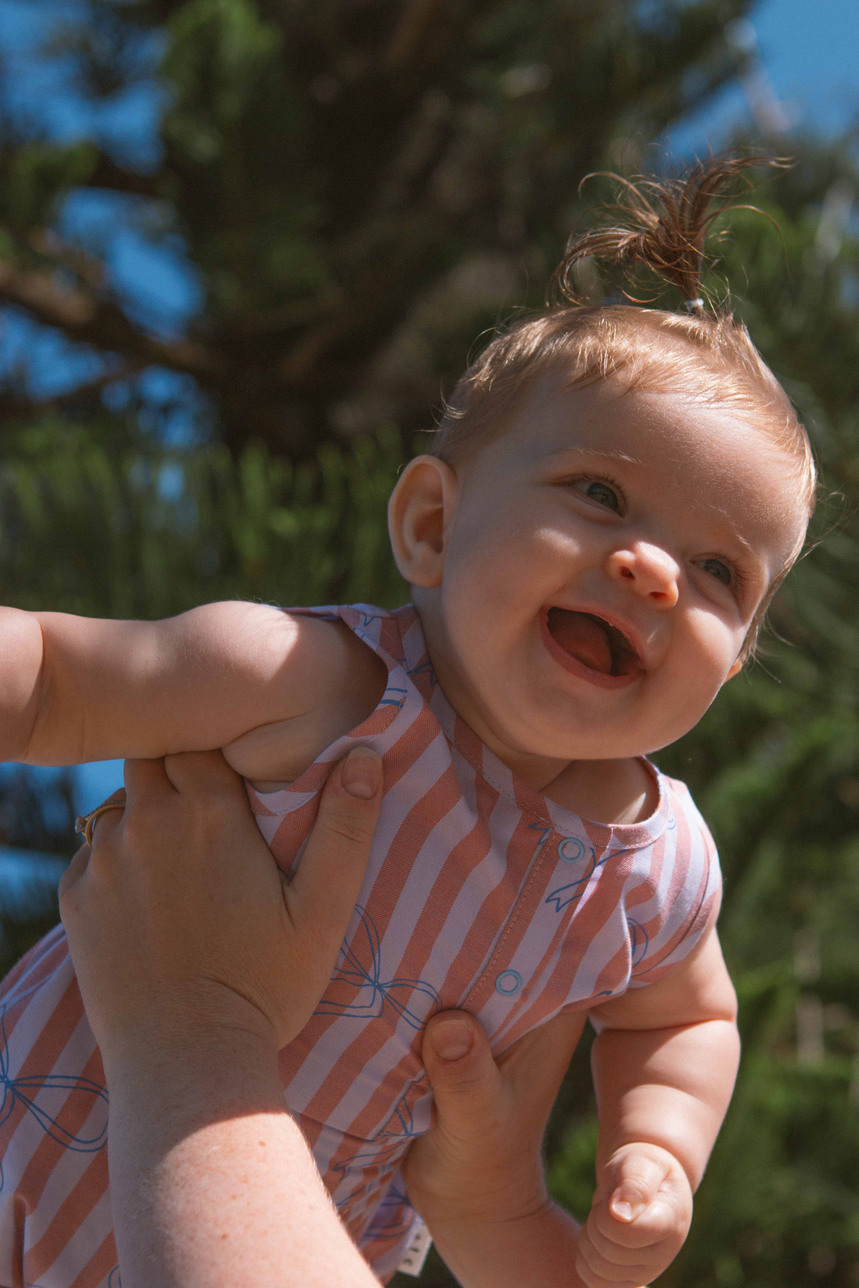 A smiling baby in a sleeveless Blue Bows Bubble Romper by OLIVE + THE CAPTAIN, known for its versatile style and easy dressing, is being held up in the air by adult hands. The baby has a small tuft of hair on top of their head, and the background features green pine tree branches and a clear blue sky.