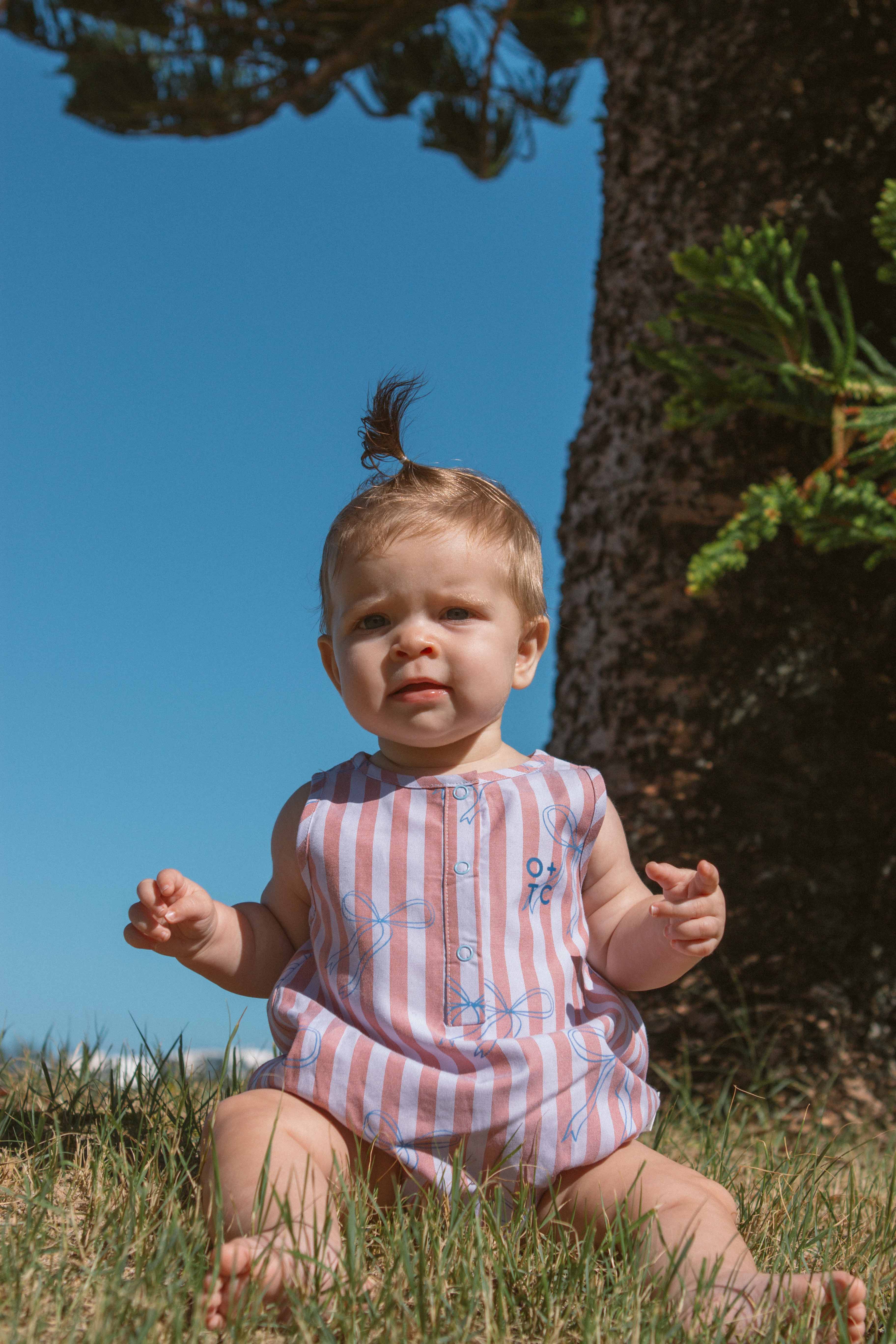 A baby sitting on the grass in front of a large tree is wearing the Blue Bows Bubble Romper by OLIVE + THE CAPTAIN. The easy dressing design of this adorable piece complements the small sprout of hair tied up on top of their head as they look towards the camera with a neutral expression. The sky in the background is clear and blue.