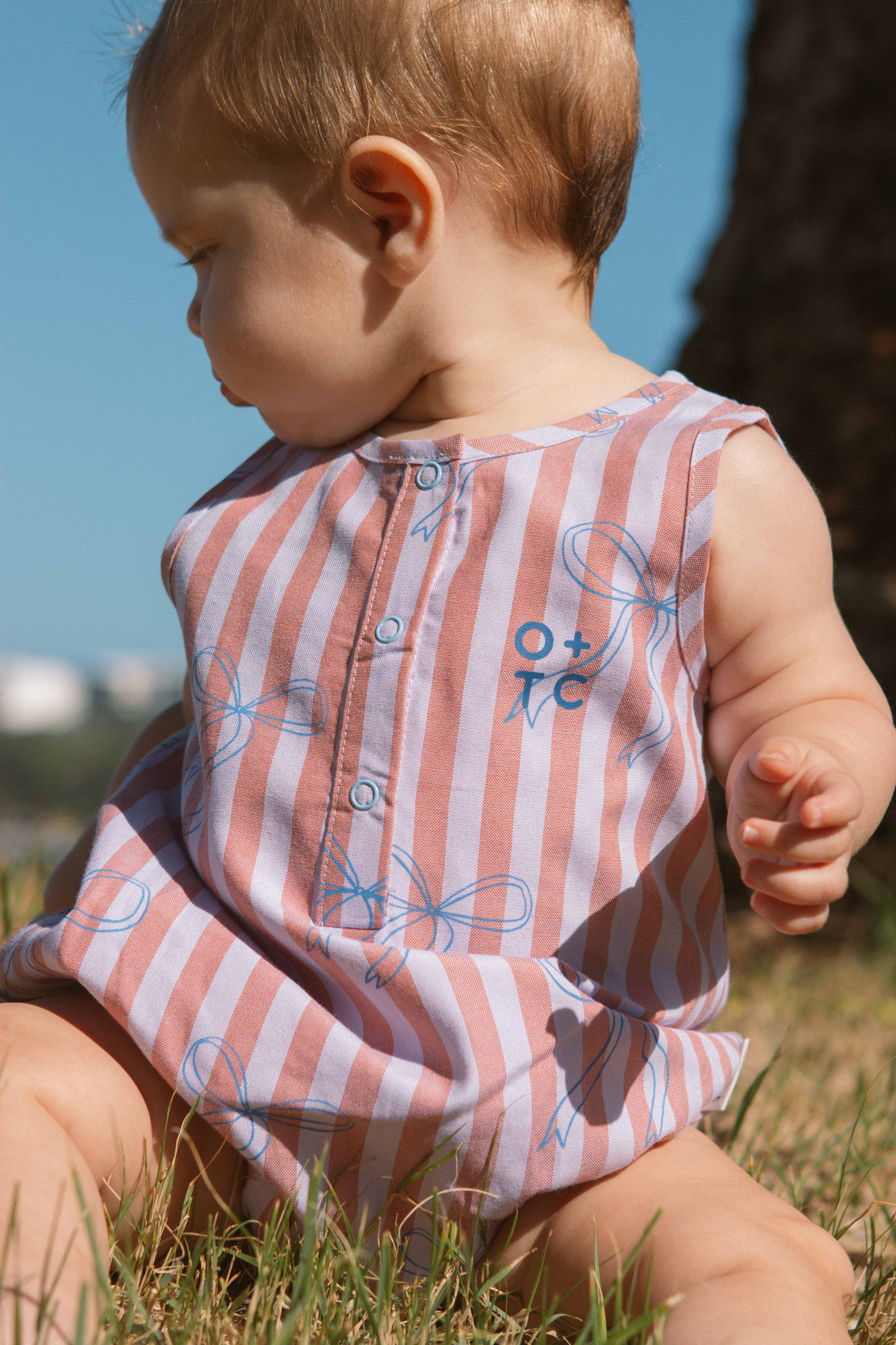 A baby sits on grass, looking to the side. The baby is wearing the Blue Bows Bubble Romper from OLIVE + THE CAPTAIN, a sleeveless, buttoned piece adorned with pink and white vertical stripes and charming blue bow illustrations, along with the letters "O+TC." With its versatile style that ensures easy dressing, this adorable romper is perfect for any sunny outdoor adventure.