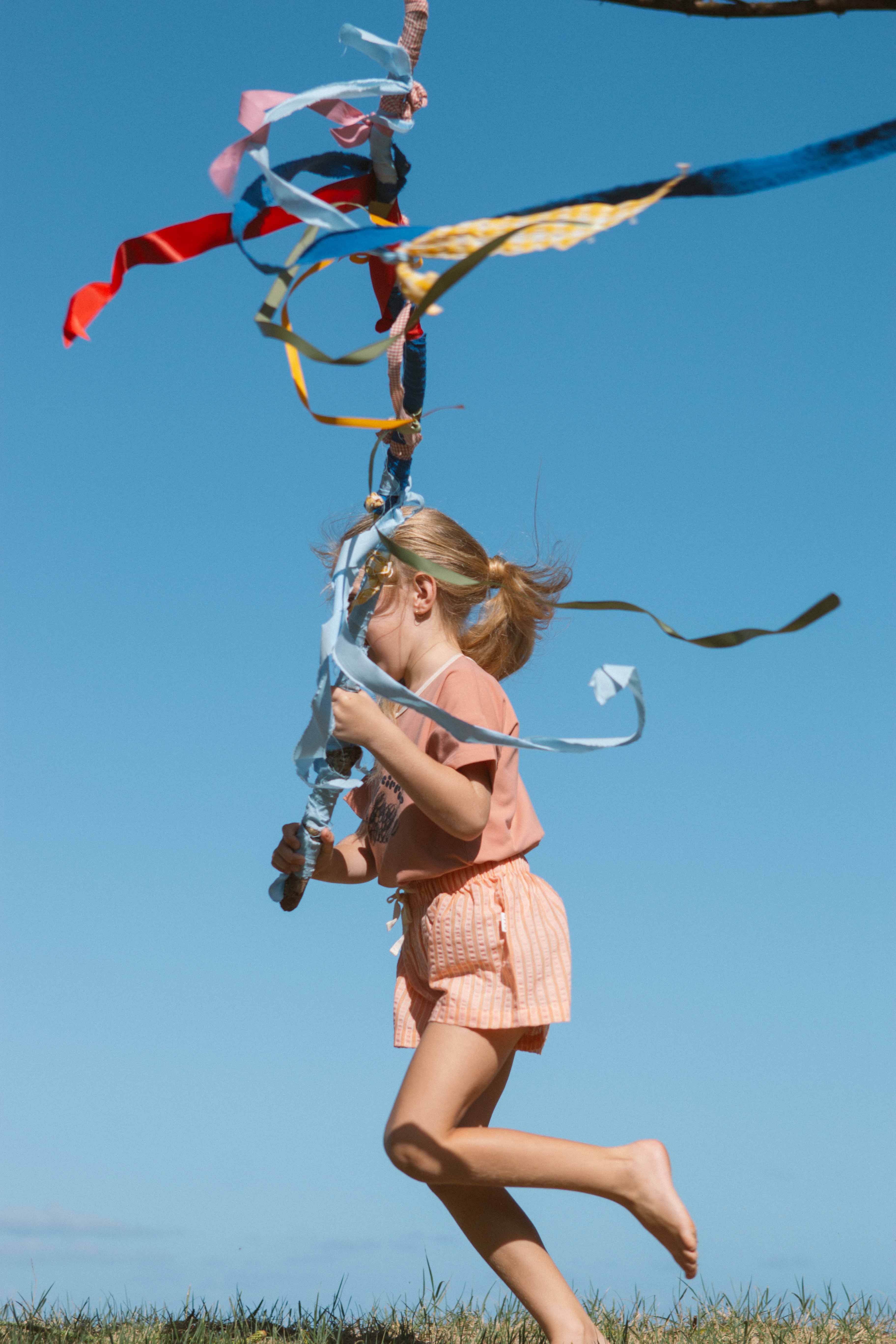 A young girl with blond hair in a ponytail is running barefoot on the grass under a clear blue sky. She is holding a stick adorned with colorful ribbons streaming in the wind. She is wearing a peach-colored shirt and OLIVE + THE CAPTAIN's Cantaloupe Wide Shorts, which are mid-thigh length and made from premium cotton polyester blend.