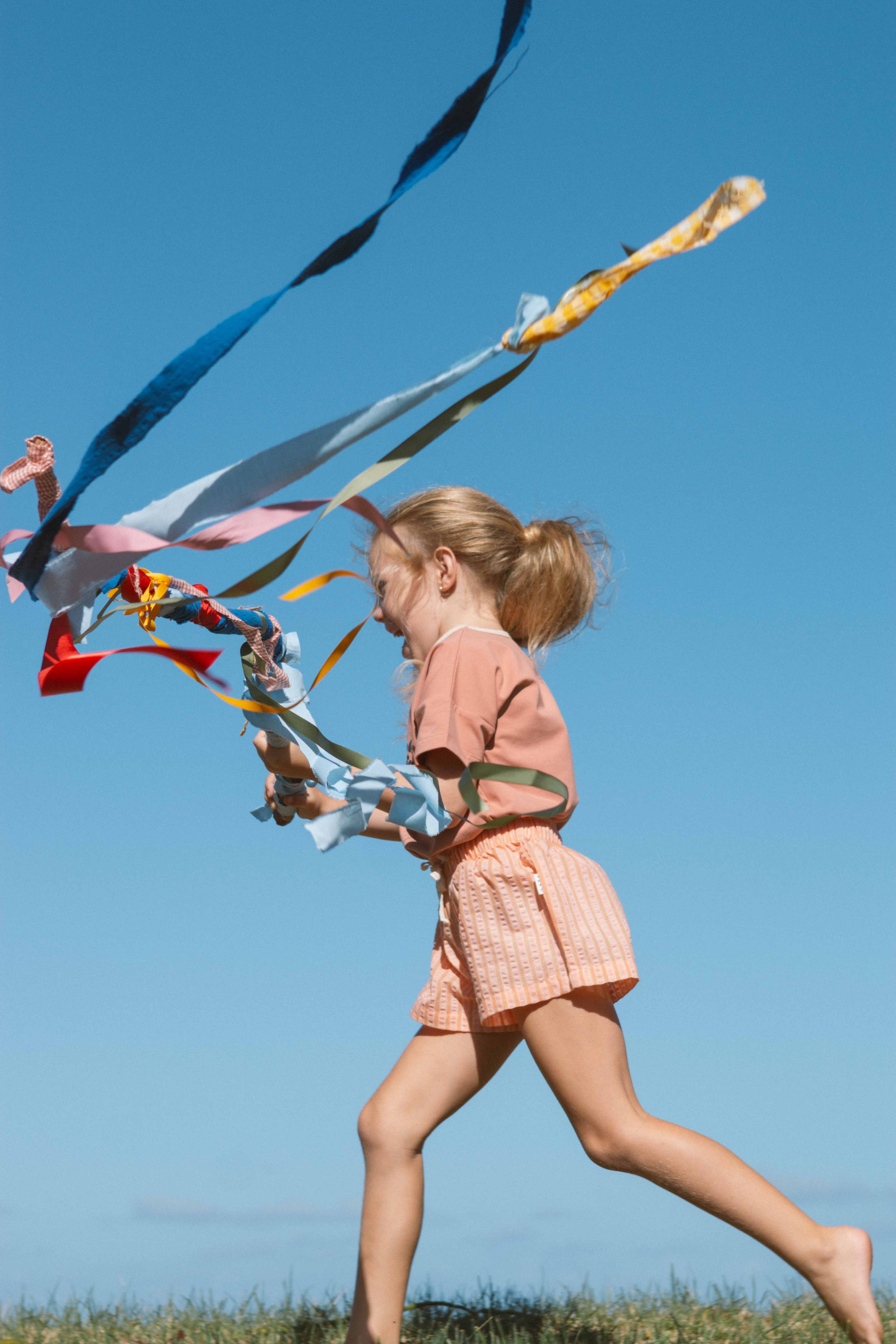 A young girl with blonde hair in a ponytail is joyfully running barefoot in a grassy field. She is wearing a pink shirt and Cantaloupe Wide Shorts made of premium cotton polyester blend from OLIVE + THE CAPTAIN, and is holding long, colorful streamers that flutter in the wind against a clear blue sky.