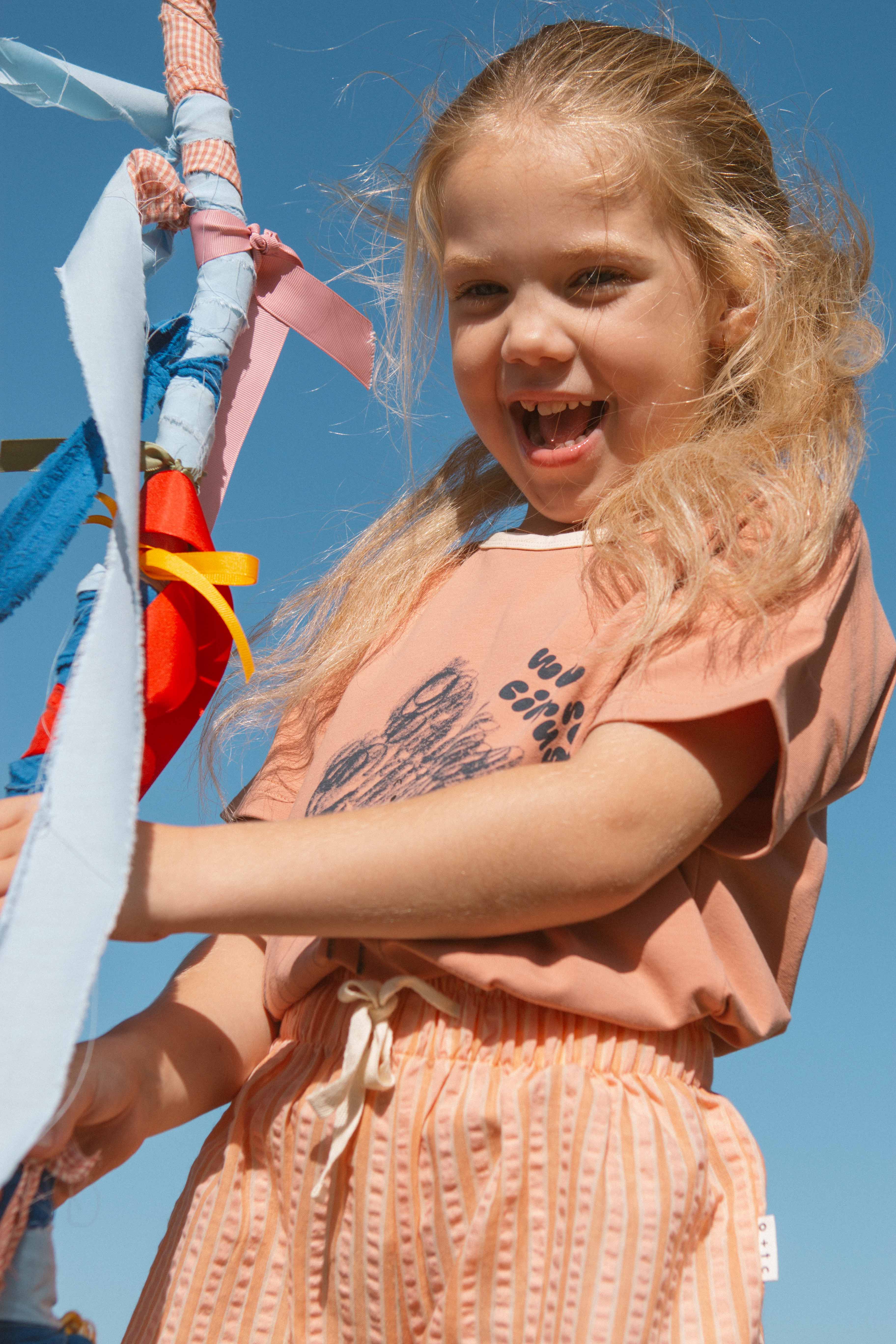 A young girl with blonde hair smiles joyfully, standing outdoors against a clear blue sky. She is wearing a peach-colored shirt and the Cantaloupe Wide Shorts from OLIVE + THE CAPTAIN, made from a premium cotton polyester blend, and is holding onto colorful ribbons wrapped around a pole.