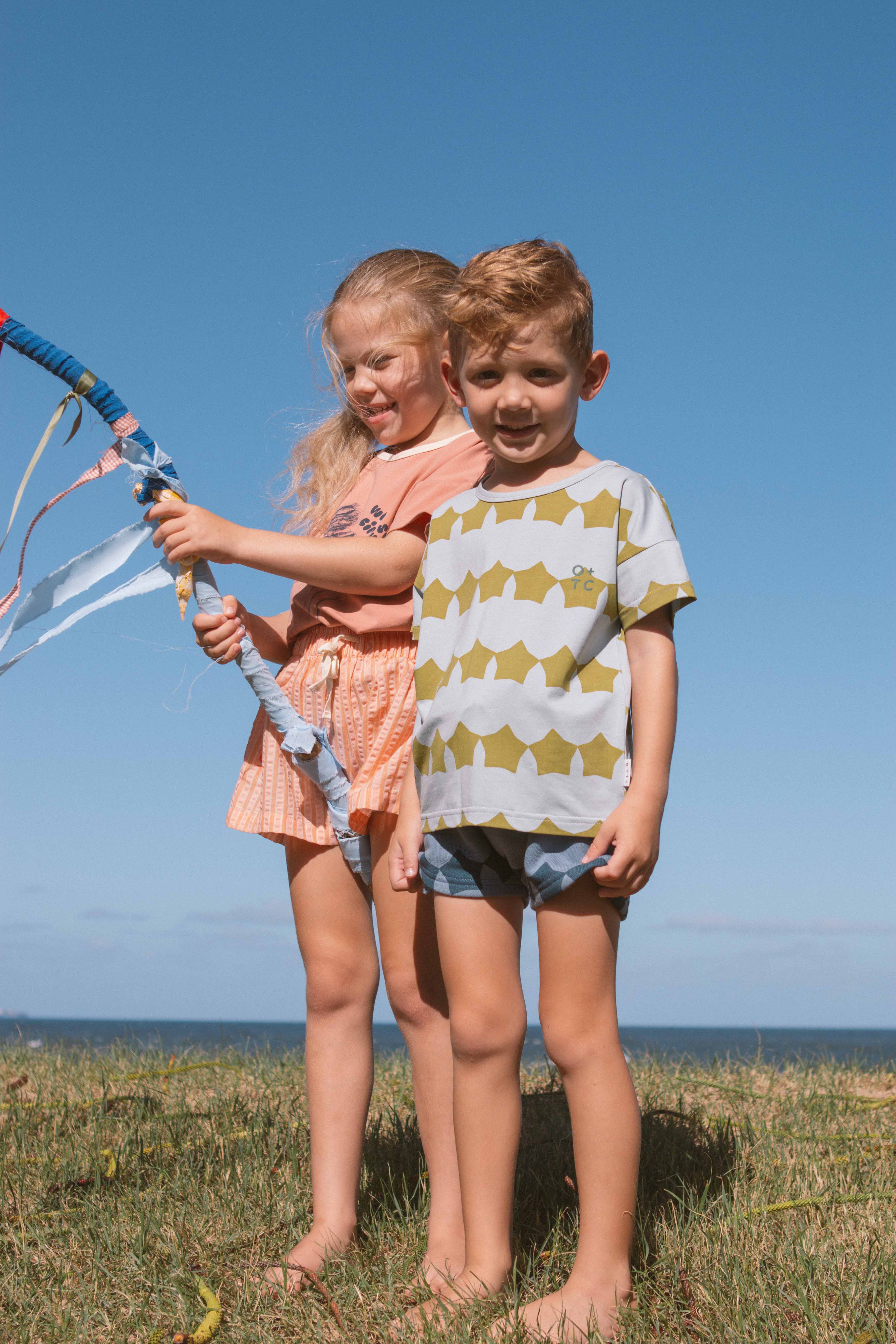 Two children standing on grass near the ocean. The girl on the left is holding a colorful kite and wearing a peach-colored outfit made of super soft fabric, while the boy on the right is wearing OLIVE + THE CAPTAIN's Evergreen Supernova Relaxed Fit Tee in green and white stripes paired with blue shorts, designed in an elevated classic style. The sky is clear and sunny.