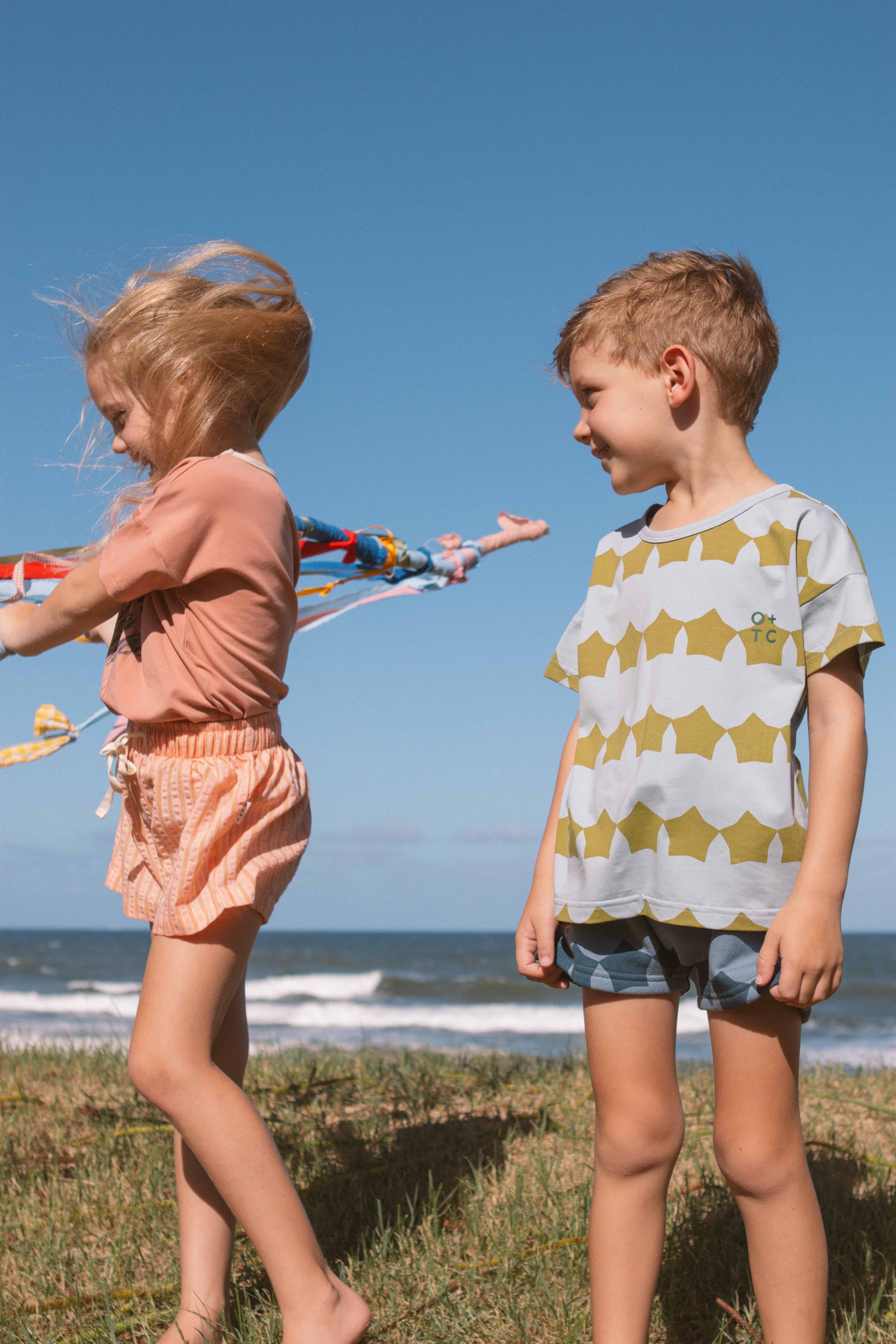 Two children are playing near the beach. One child with long hair, dressed in an Evergreen Supernova Relaxed Fit Tee from OLIVE + THE CAPTAIN made of premium cotton, is flying a colorful kite, while the other child, with short hair and also wearing an Evergreen Supernova Relaxed Fit Tee from OLIVE + THE CAPTAIN made of super soft fabric, watches and smiles. The sky is clear and blue, and the ocean waves can be seen in the background.