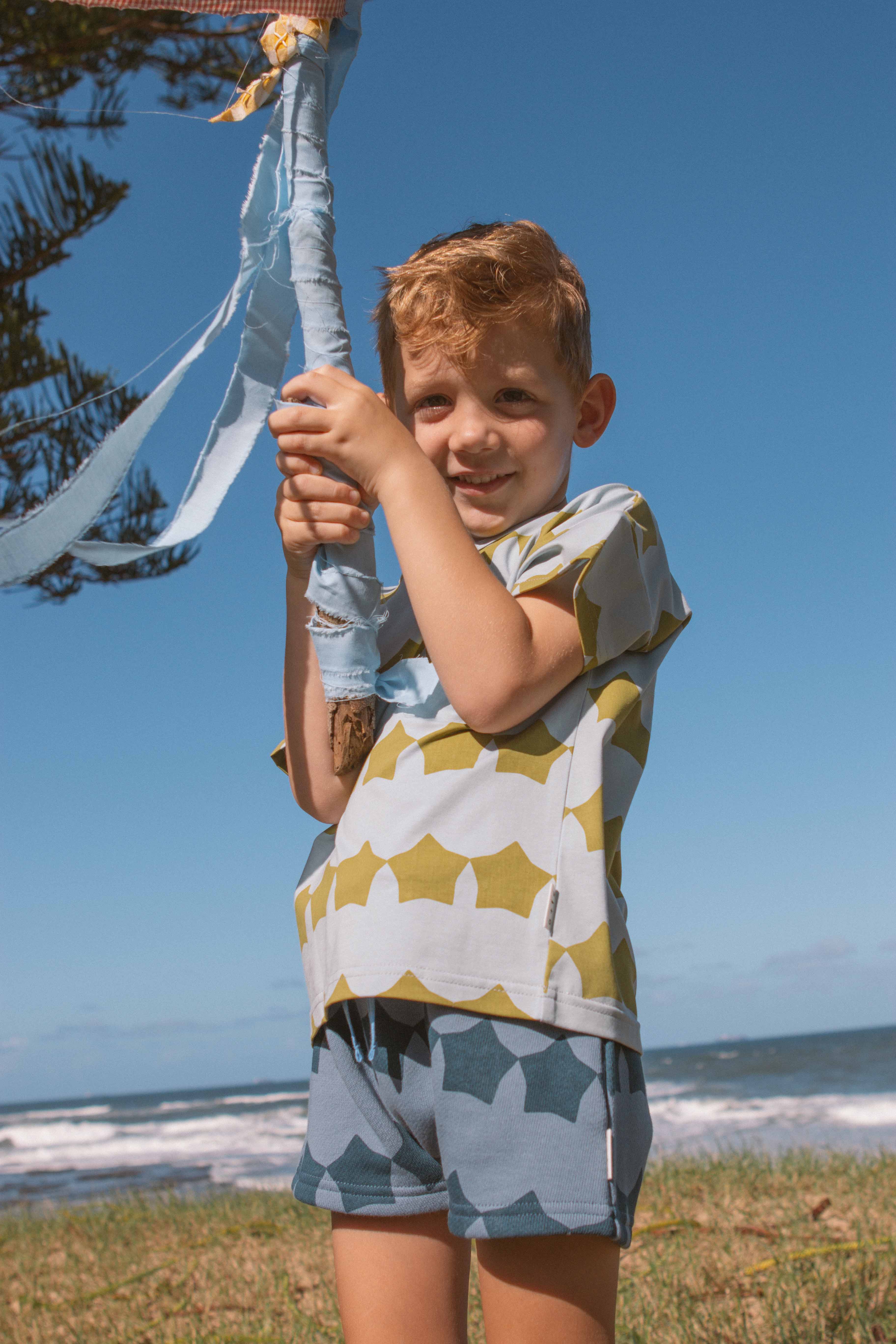 A young boy with blonde hair is wearing the Midnight Supernova Shorties, made from 100% cotton by OLIVE + THE CAPTAIN, and smiles while holding onto a fabric-wrapped pole on a grassy hill. The backdrop features a sunny seaside view with waves crashing onto the shore under a clear blue sky.