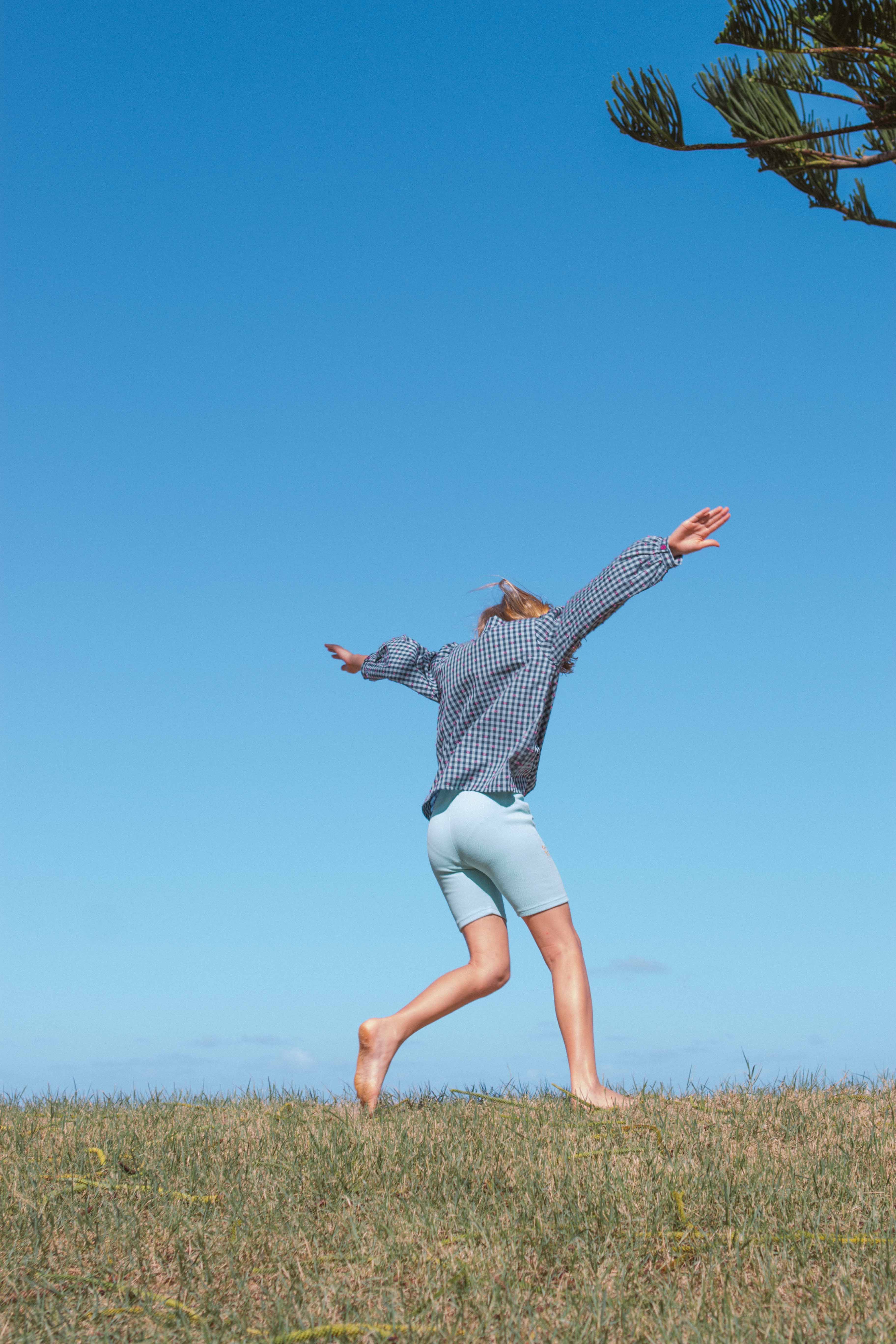 A person wearing a blue checkered shirt and OLIVE + THE CAPTAIN's Aqua Sky Bike Shorts is captured mid-leap in an open grassy field against a clear blue sky. They are barefoot, with arms outstretched, and a tree branch is visible in the upper right corner of the image.