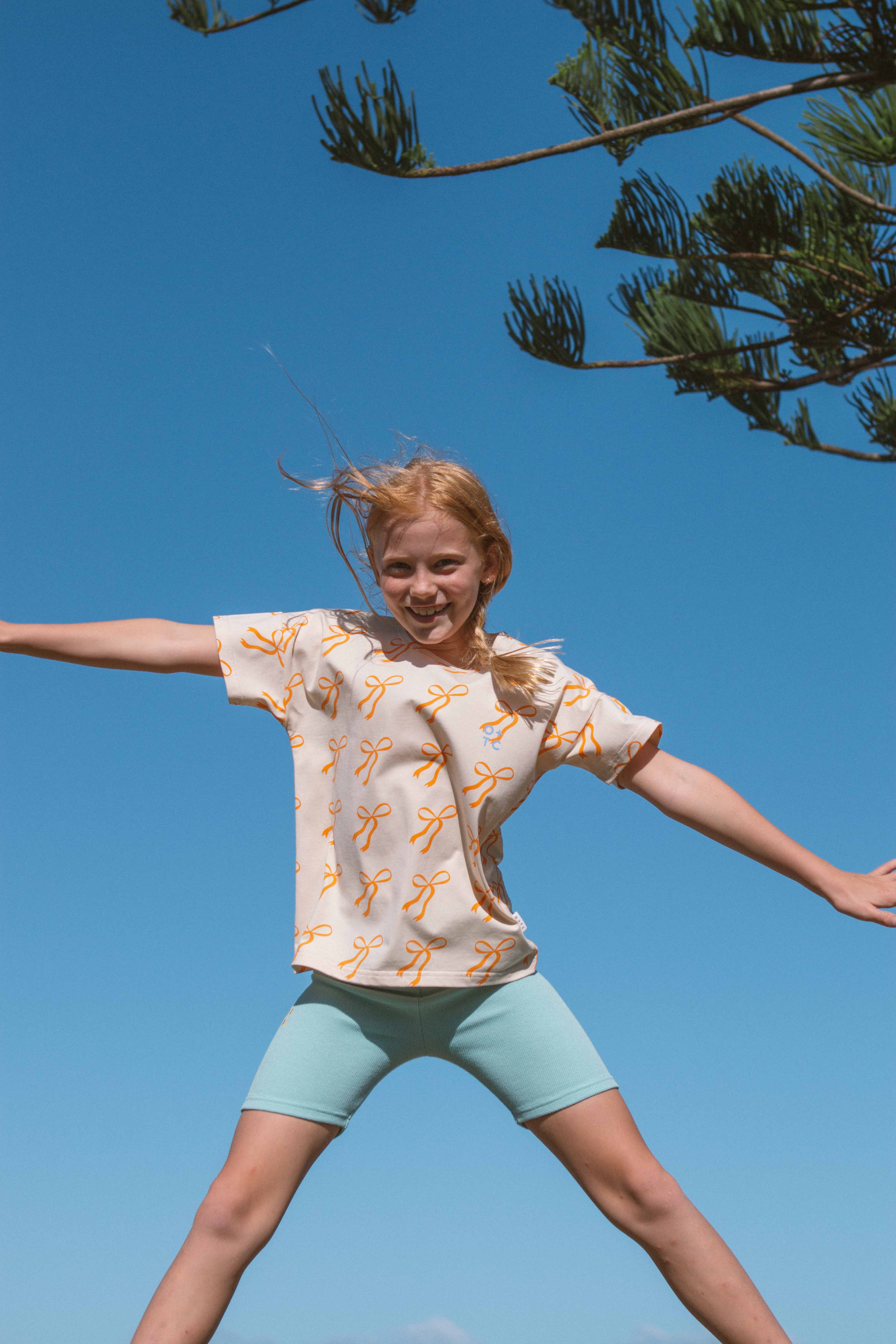 A girl with blonde hair is jumping joyfully against a clear blue sky, her Cream Bows Relaxed Fit Tee from OLIVE + THE CAPTAIN and light green shorts catching the sunlight. The soft jersey of her top, in a boxy fit, adds to the carefree vibe as a tree branch with green needles extends into the frame from the right side.