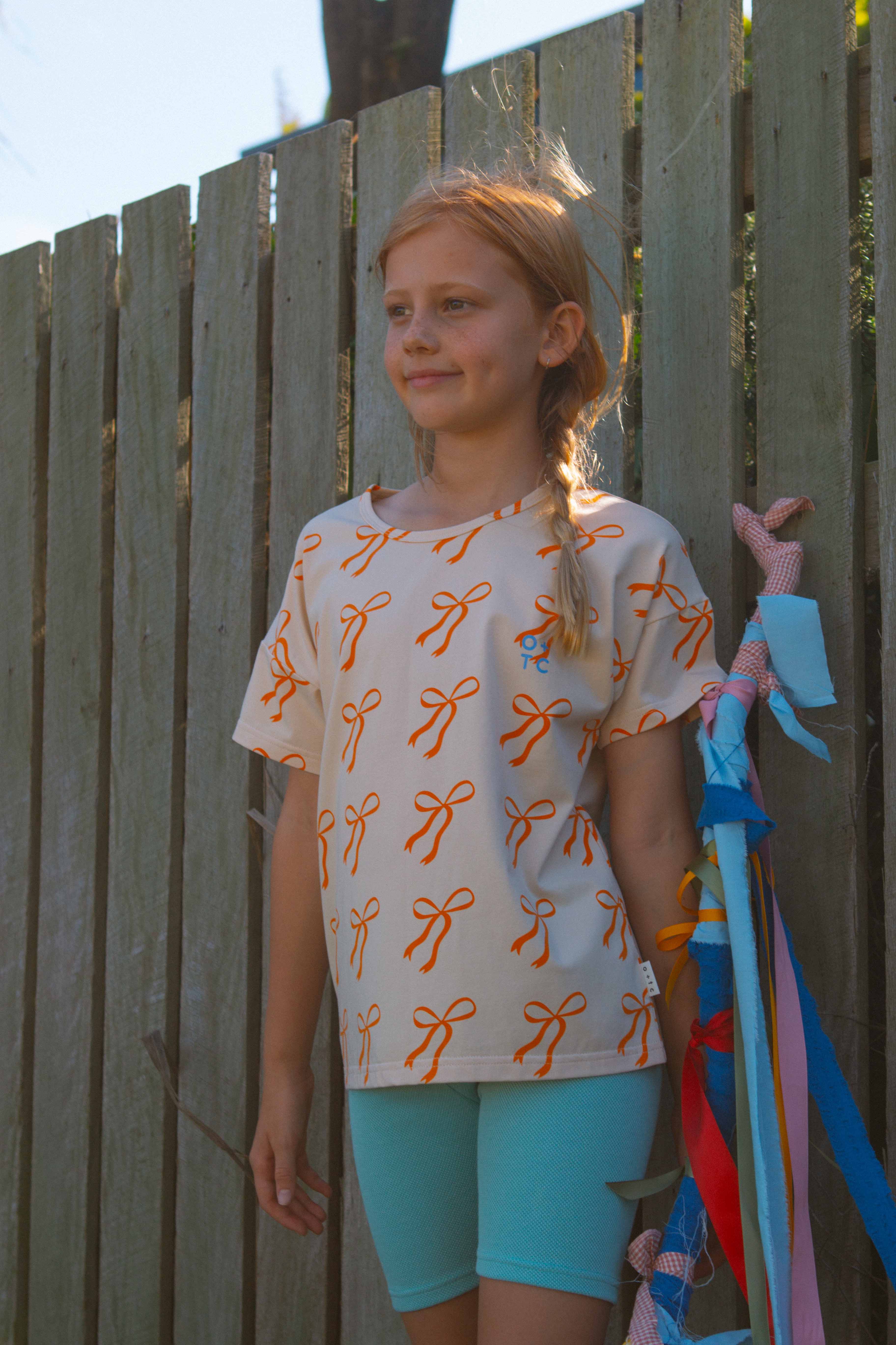 A young girl stands in front of a wooden fence, wearing the OLIVE + THE CAPTAIN Cream Bows Relaxed Fit Tee paired with teal shorts. Her premium cotton blend top, featuring an orange bow pattern, boasts a boxy fit that enhances her casual charm. She holds a bundle of colorful ribbons in her right hand and has a small smile on her face. Her hair is styled in a side braid.