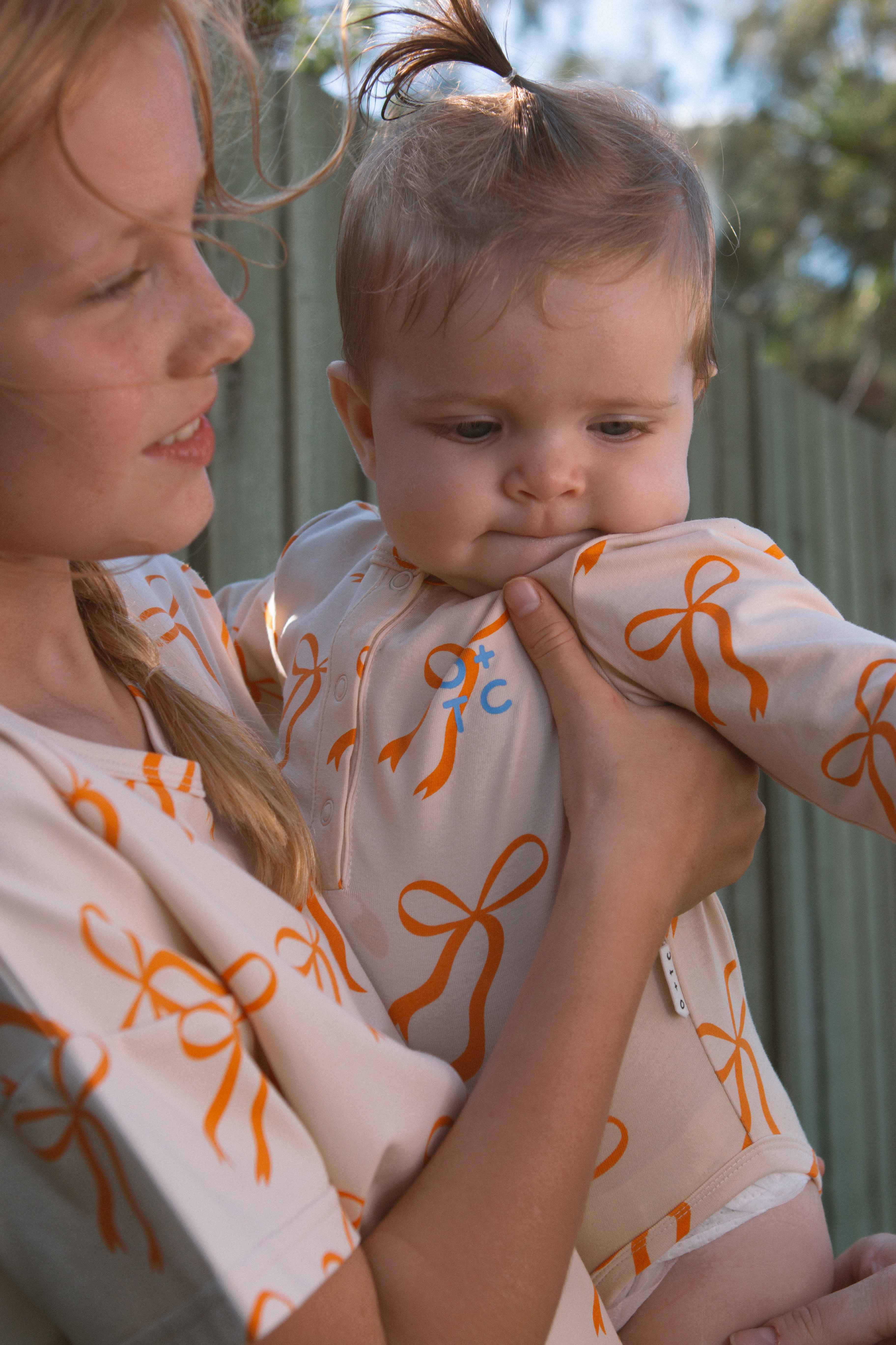 A woman is holding a baby outdoors on a sunny day. Both are wearing matching outfits with golden bows. The woman is smiling and looking at the baby, who is wearing an OLIVE + THE CAPTAIN Cream Bows Long Sleeve Bodysuit and gazing off to the side. A green fence and trees are visible in the background.