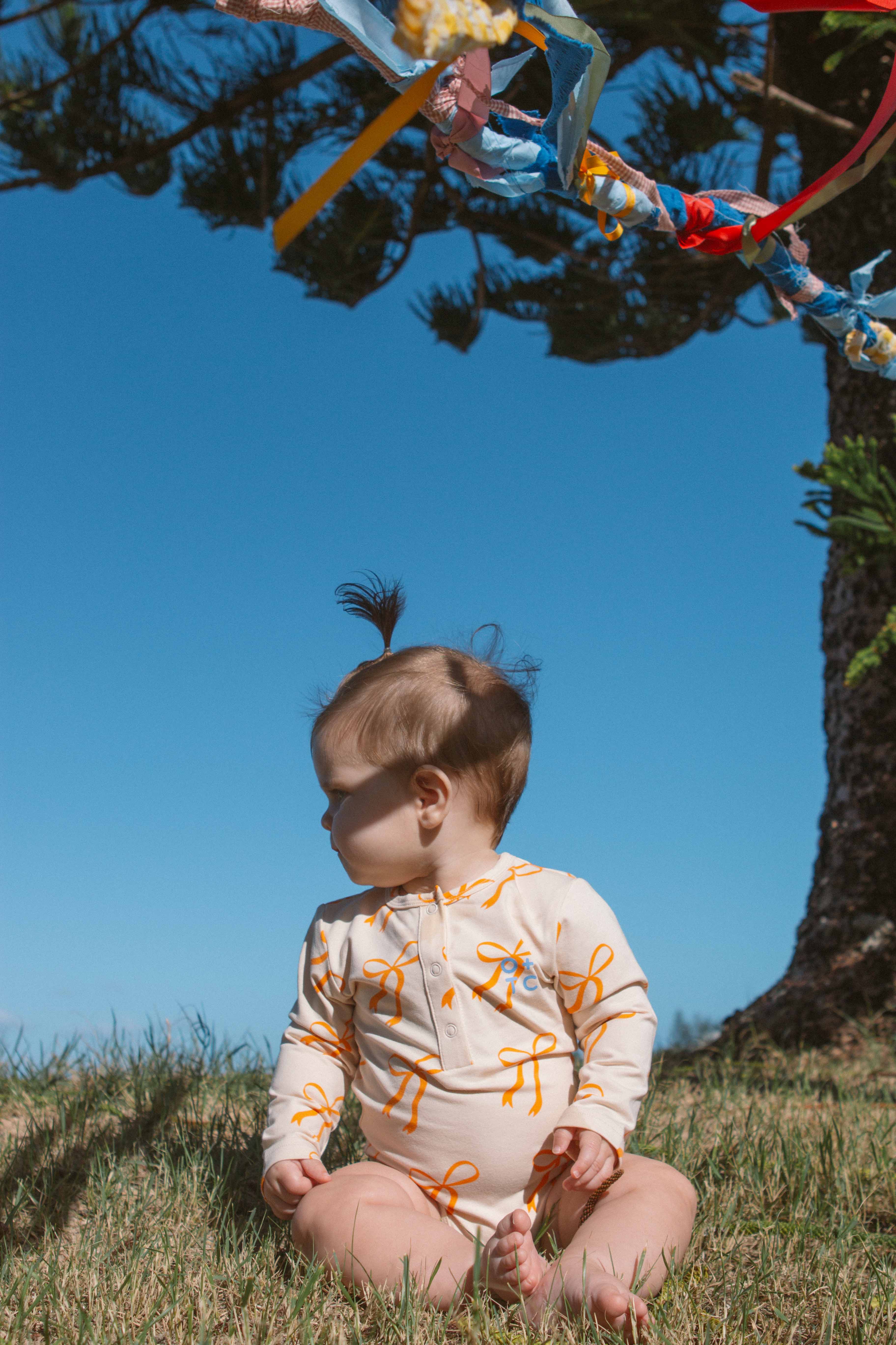 A baby sits on a grassy field under a clear blue sky, turned slightly to the side. The baby wears OLIVE + THE CAPTAIN's Cream Bows Long Sleeve Bodysuit, decorated with light-colored orange patterns and has a tuft of hair tied up on top of their head. Golden bows and colorful ribbons are tied to a nearby tree branch.