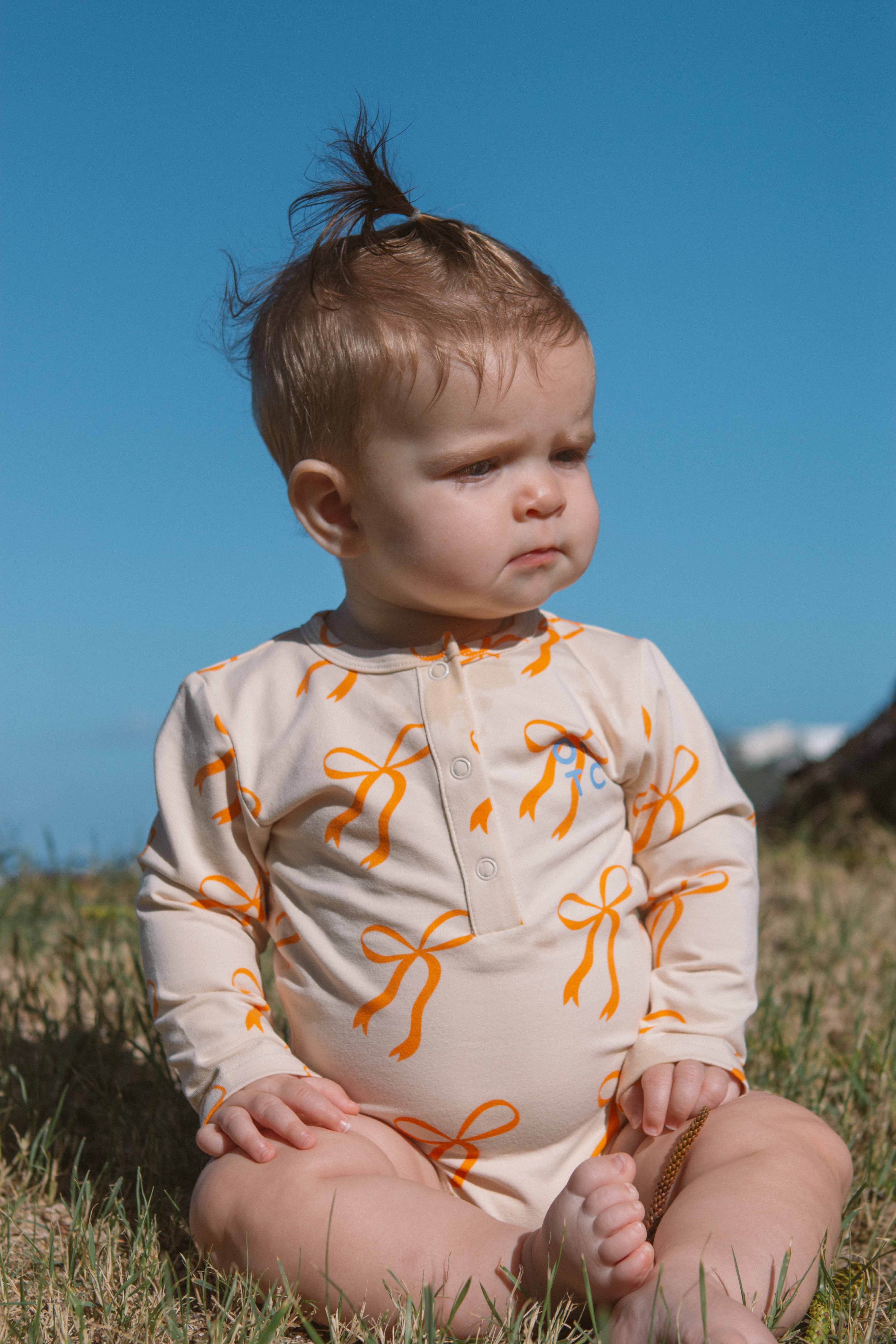 On a bright, sunny day, a baby sits on the grass under a clear blue sky. The baby is dressed in an OLIVE + THE CAPTAIN Cream Bows Long Sleeve Bodysuit and has short, light brown hair with a small tuft tied up on top. They appear to be thoughtful or focused on something in the distance.