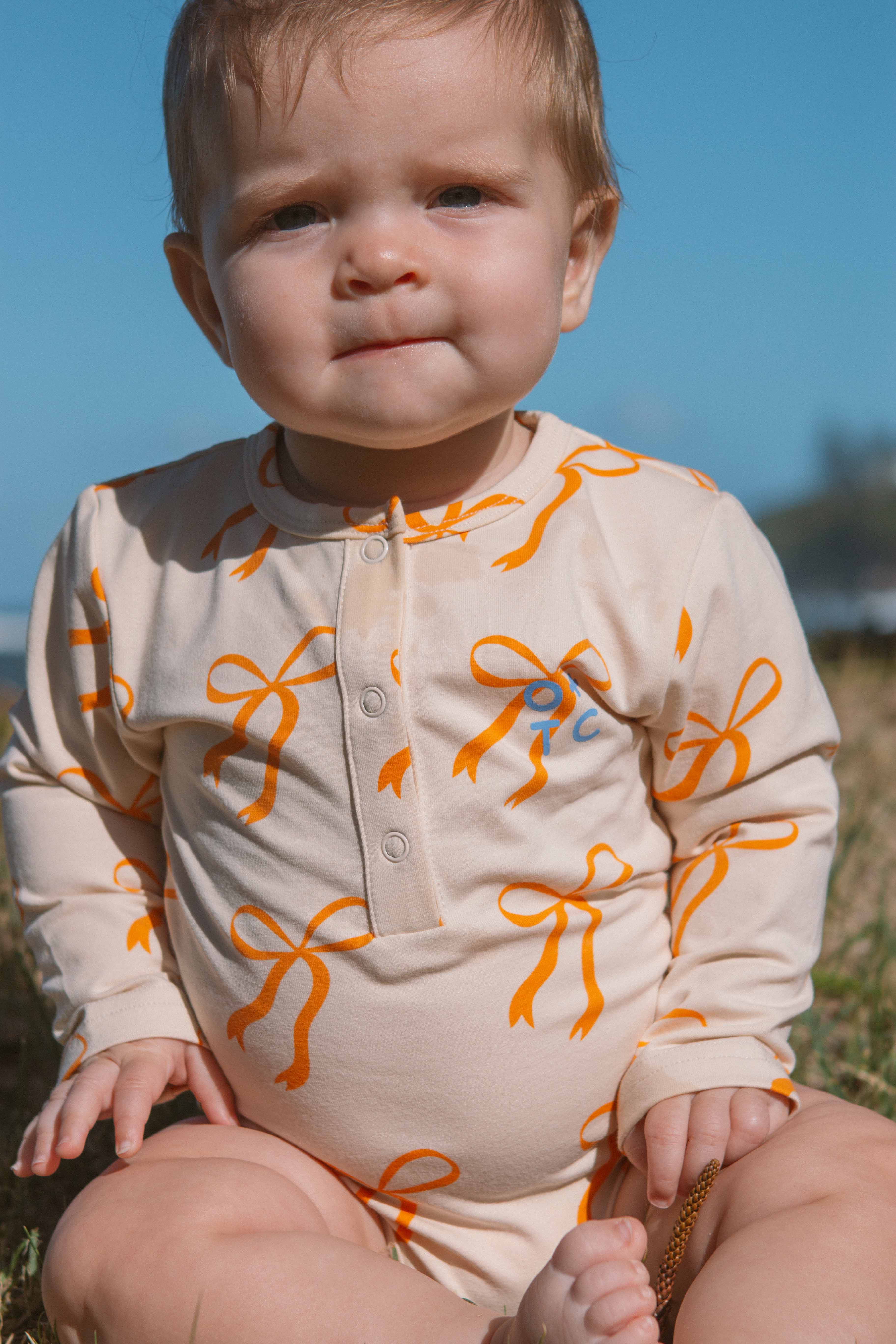 A baby sits outdoors on grass, wearing the OLIVE + THE CAPTAIN Cream Bows Long Sleeve Bodysuit. The bodysuit is adorned with golden bows. The baby has a thoughtful expression and some sunlight is visible in the background.