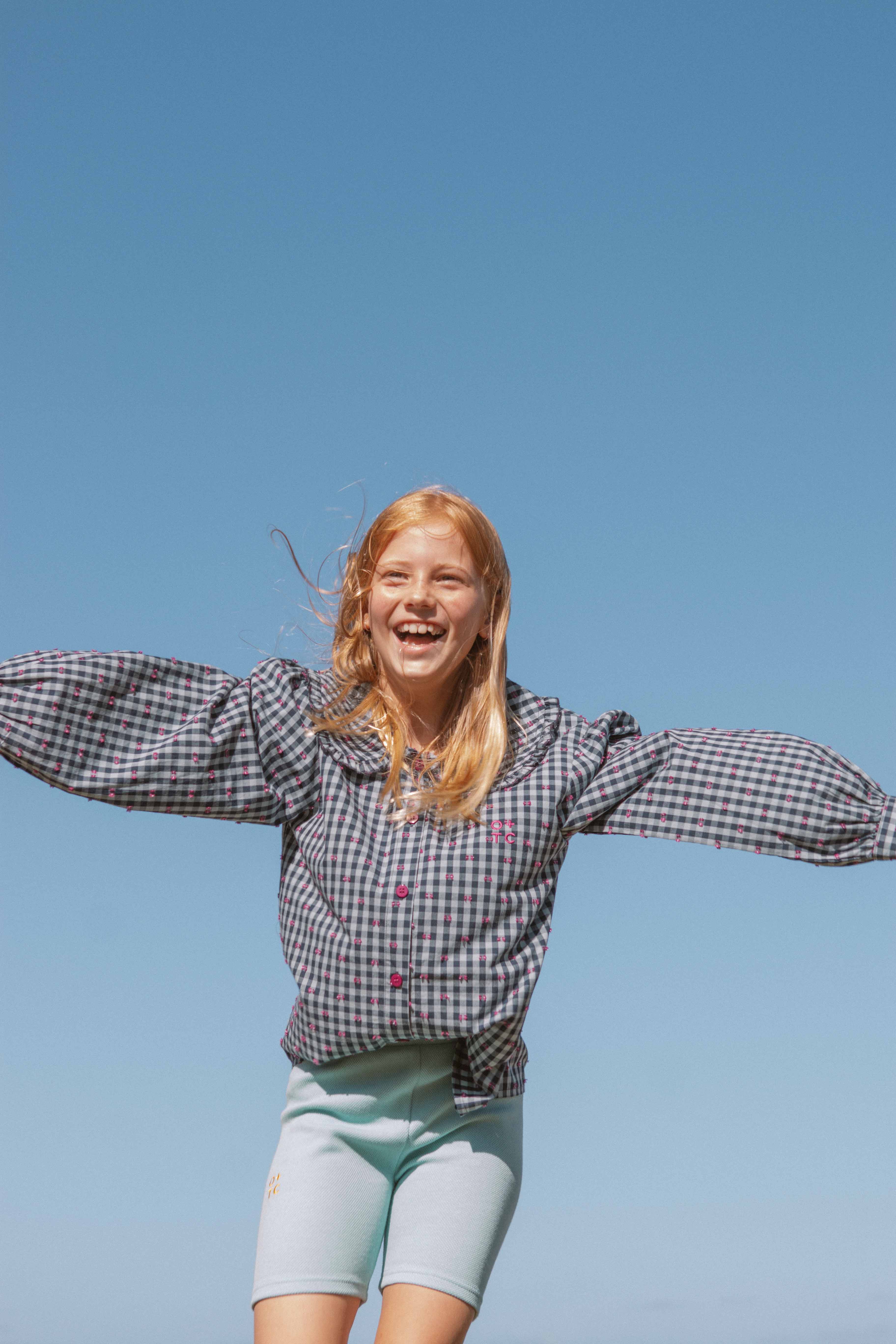 A young girl with blonde hair is joyfully jumping against a clear blue sky. She is wearing a checkered long-sleeve shirt made of premium fabric and light blue Aqua Sky Bike Shorts from OLIVE + THE CAPTAIN, featuring an elastic waist. She has a big smile on her face, and her arms are outstretched.