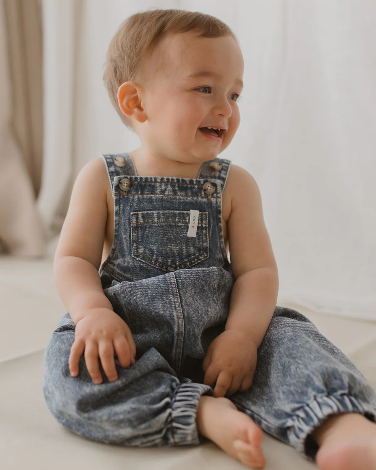 A smiling toddler wearing SUSUKOSHI's Denim Overalls Sky Blue, made from organic cotton, sits on the floor. The child, with short light brown hair, is looking to the left. The background features softly lit, light-colored flooring and neutral-draped curtains. The overalls have adjustable shoulder straps for comfort.